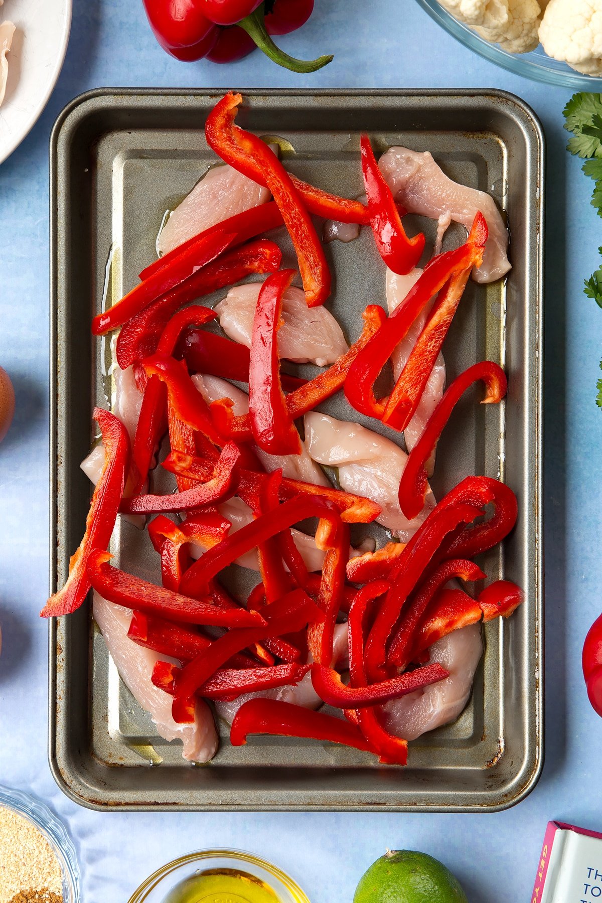 Sliced chicken breast and sliced red peppers on an oiled tray. The tray is surrounded by ingredients for cauliflower tacos. 