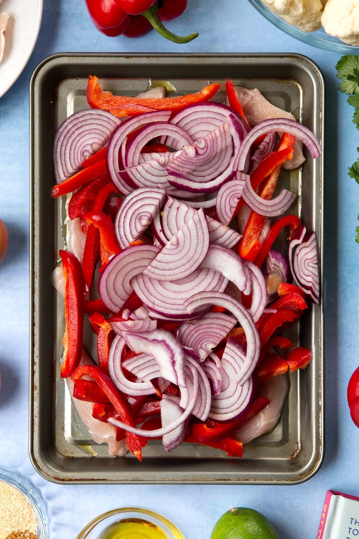 Sliced chicken breast, sliced red peppers and sliced red onion on an oiled tray. The tray is surrounded by ingredients for cauliflower tacos. 