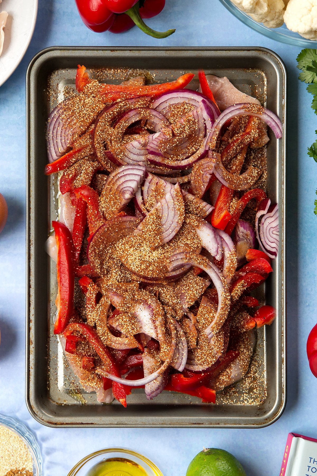 Sliced chicken breast, sliced red peppers, sliced red onion and spices on an oiled tray. The tray is surrounded by ingredients for cauliflower tacos. 