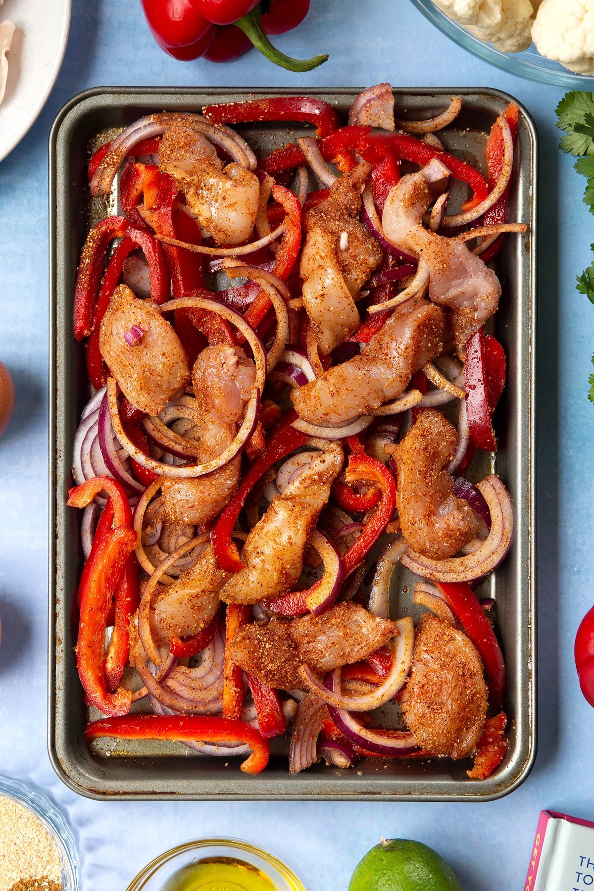 Sliced chicken breast, sliced red peppers, sliced red onion and spices mixed together on an oiled tray. The tray is surrounded by ingredients for cauliflower tacos. 