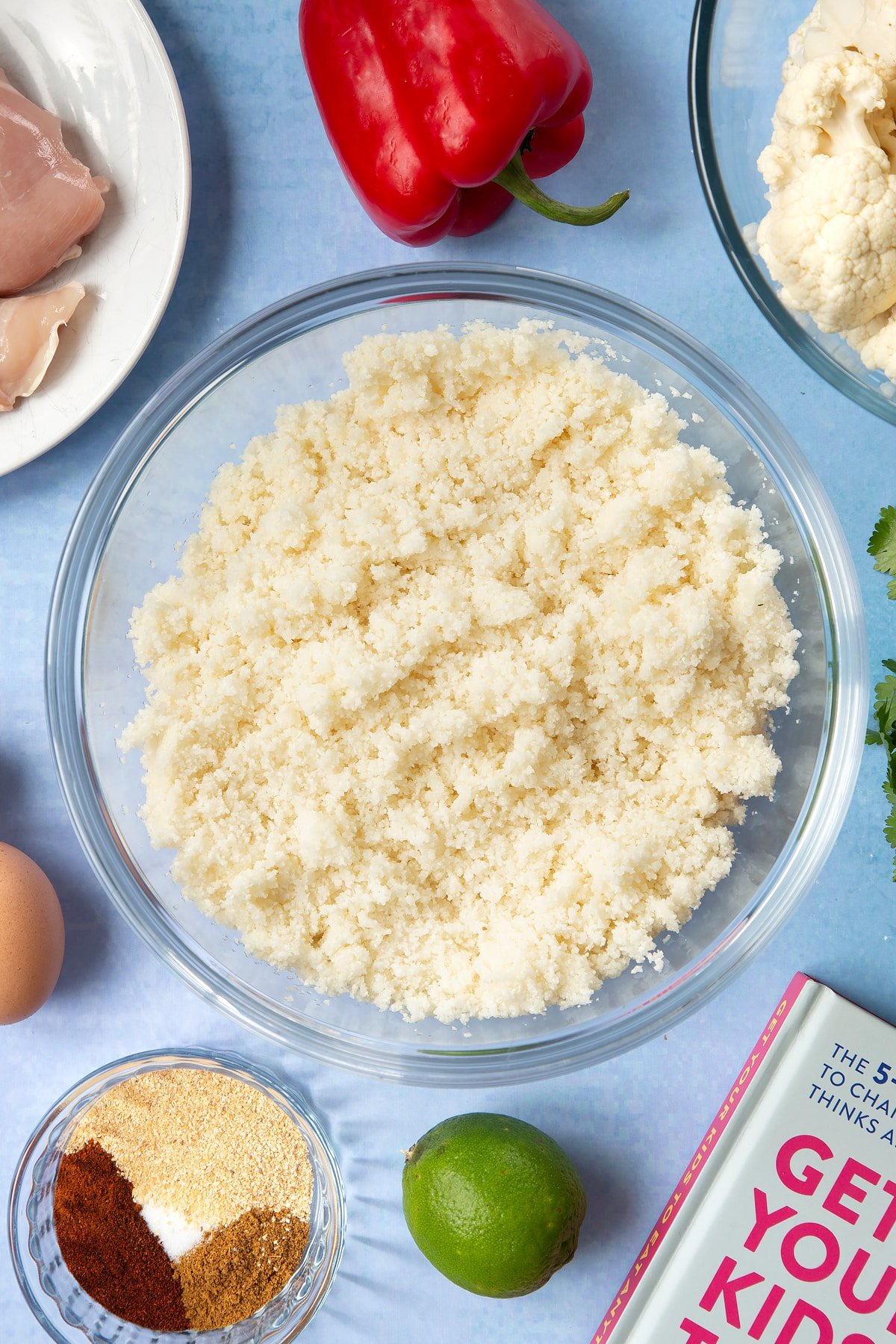 A glass mixing bowl filled with finely chopped cauliflower. The bowl is surrounded by ingredients for cauliflower tacos. 