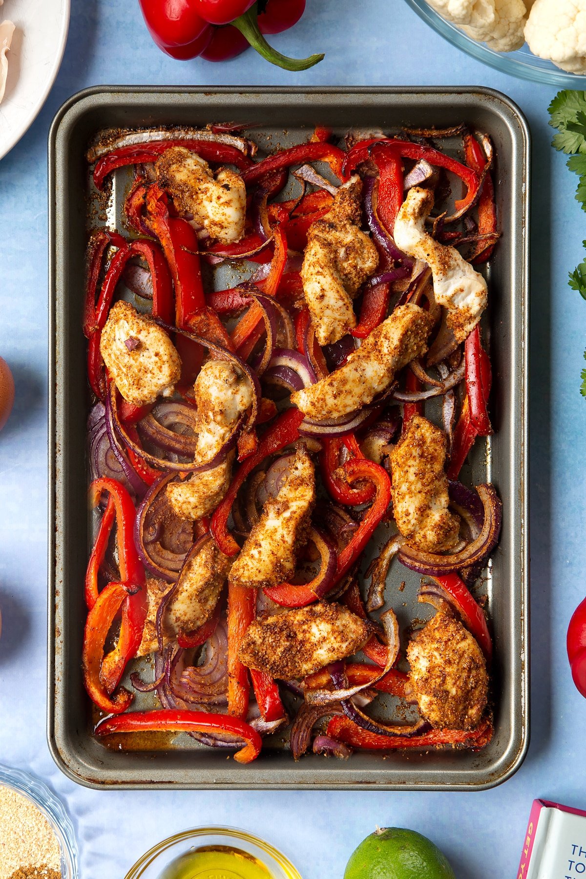 Sliced chicken breast, sliced red peppers, sliced red onion and spices mixed together on an oiled tray and roasted. The tray is surrounded by ingredients for cauliflower tacos. 