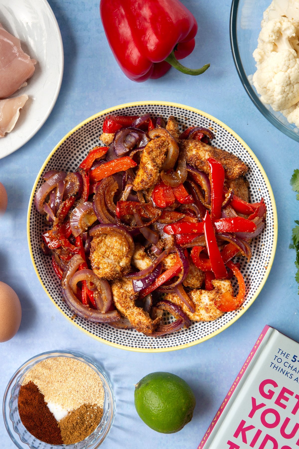 Roasted chicken breast, red peppers, red onion and spices in a bowl. The bowl is surrounded by ingredients for cauliflower tacos. 