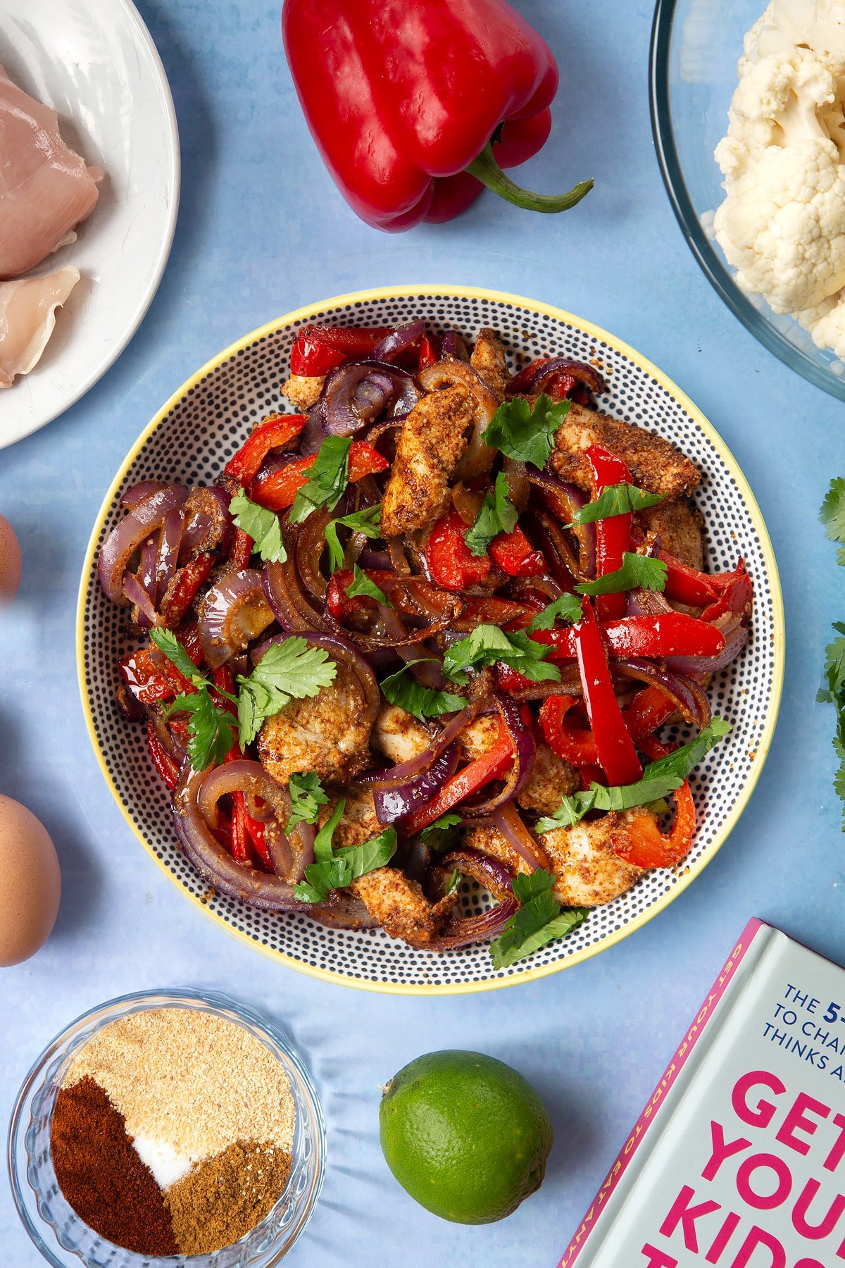 Roasted chicken breast, red peppers, red onion and spices in a bowl, topped with freshly chopped coriander. The bowl is surrounded by ingredients for cauliflower tacos. 