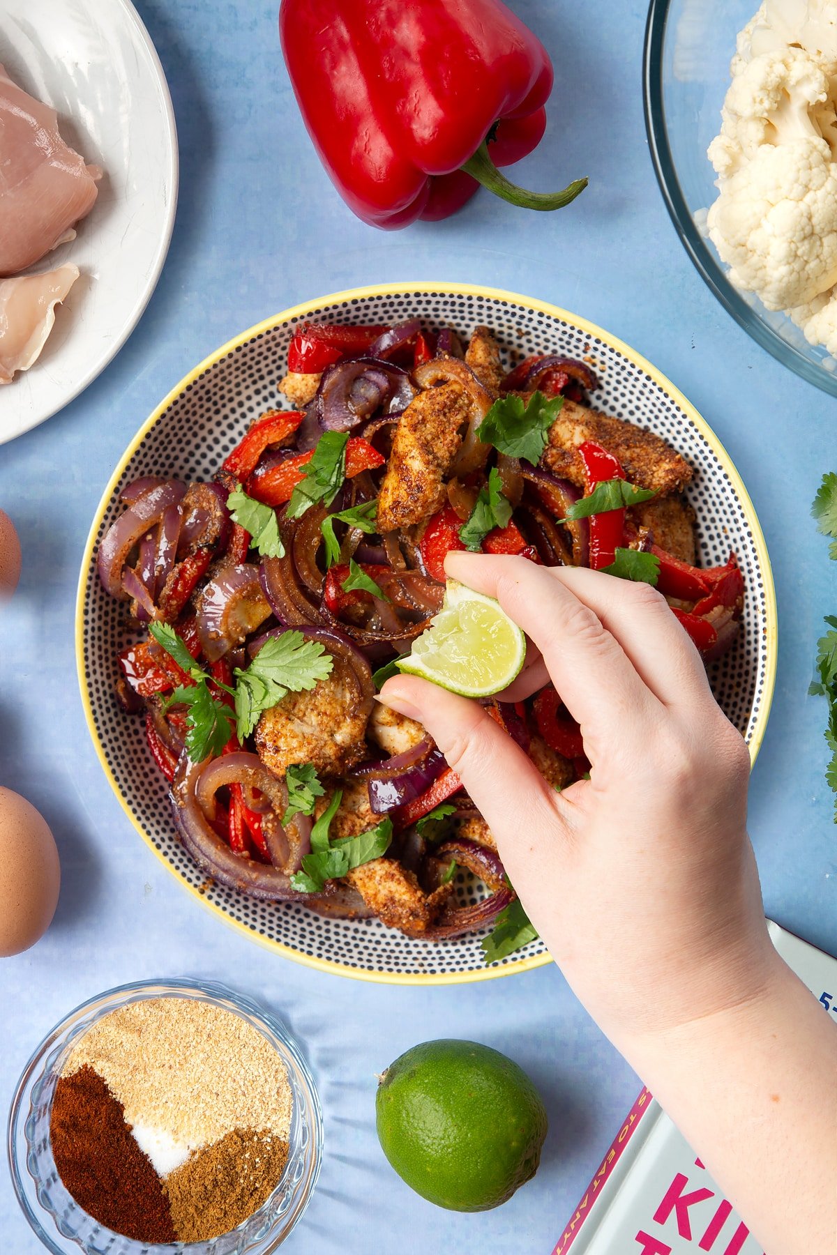 Roasted chicken breast, red peppers, red onion and spices in a bowl, topped with freshly chopped coriander. The bowl is surrounded by ingredients for cauliflower tacos. A hand squeezes fresh lime over the chicken.