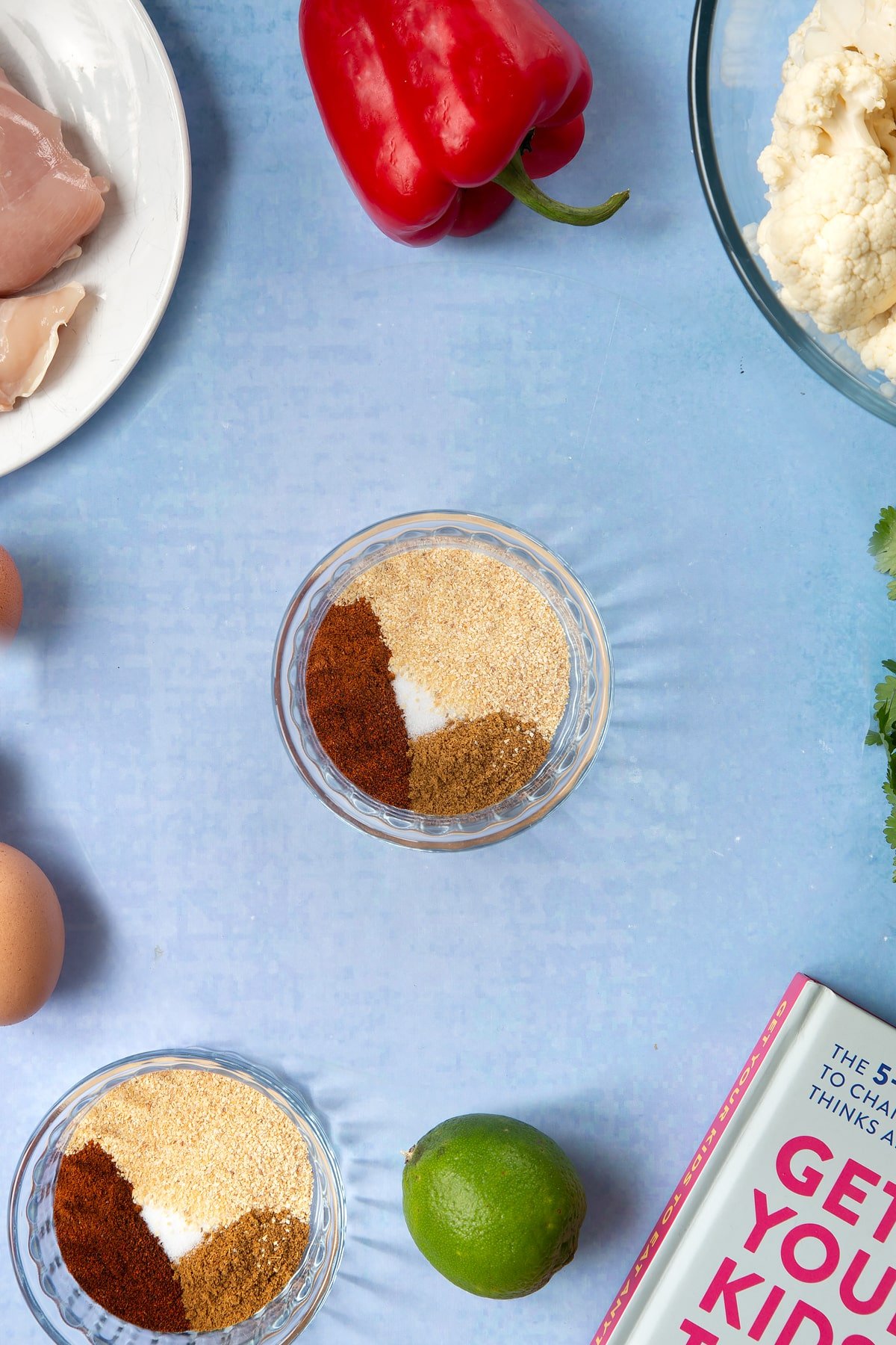 A small glass mixing bowl filled with a selection of spices. The bowl is surrounded by ingredients for cauliflower tacos. 