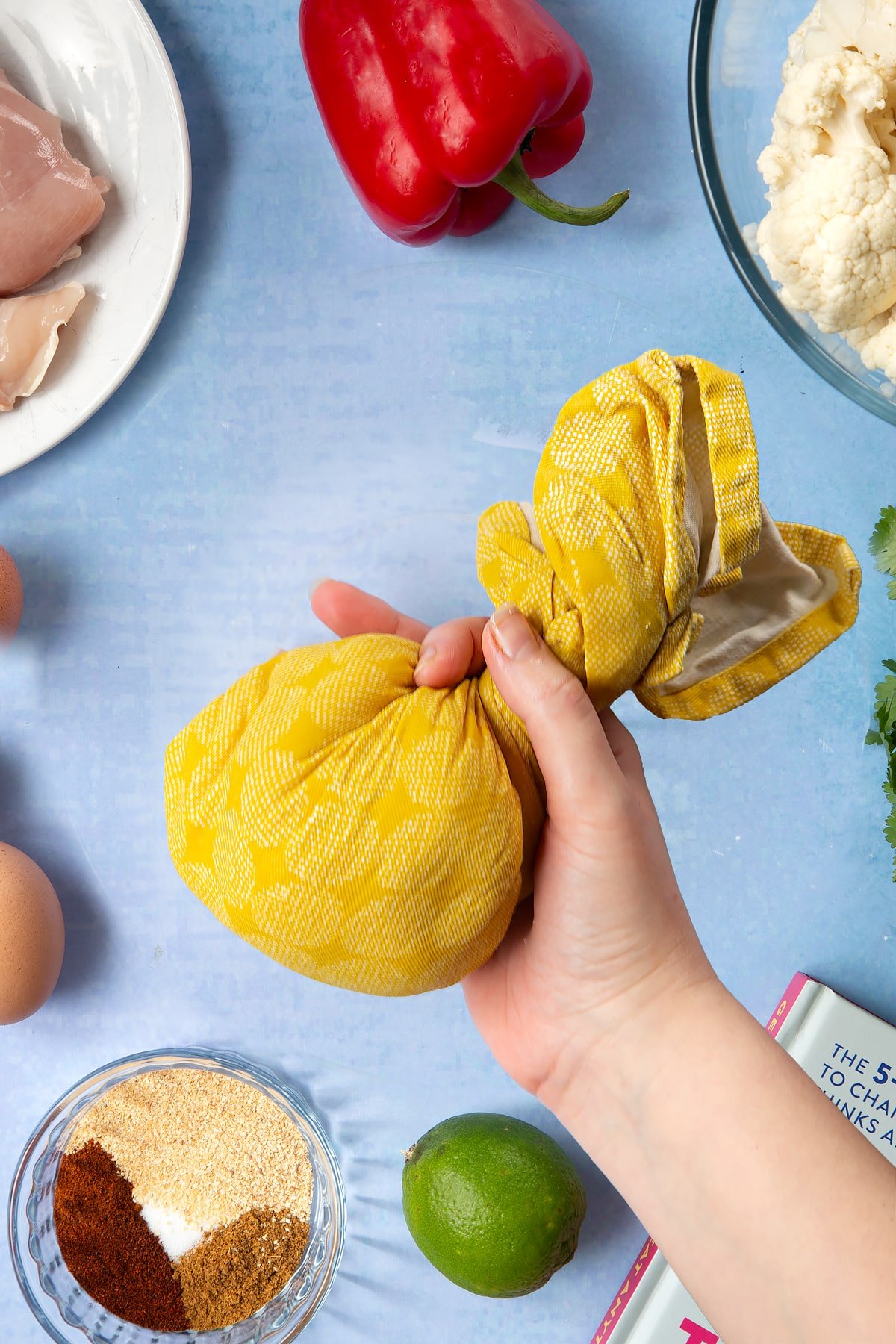 A hand holding a twisted tea towel, filled with finely chopped cauliflower. The hand is surrounded by ingredients for cauliflower tacos. 