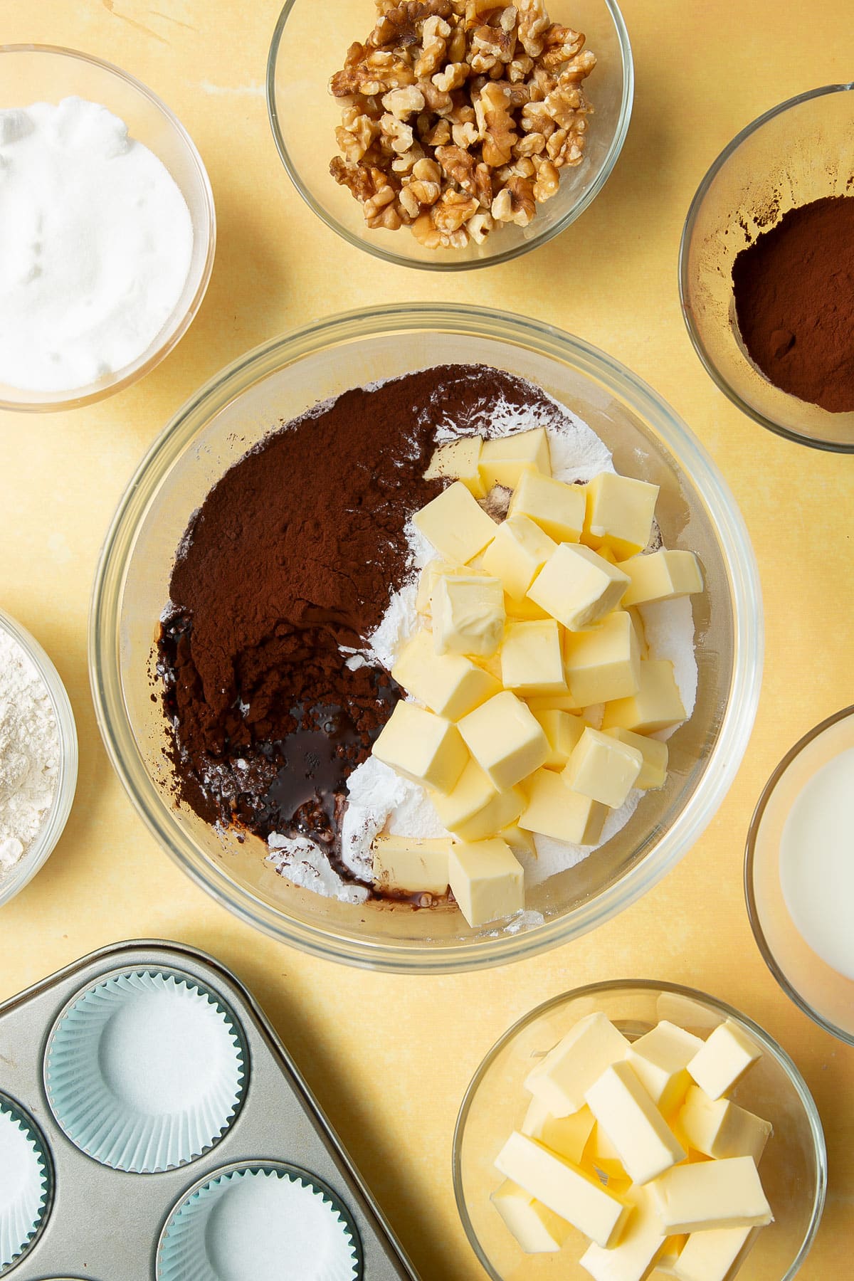 Icing sugar, butter, cocoa and hot water in a glass mixing bowl. Ingredients for chocolate walnut cupcakes surround the bowl.