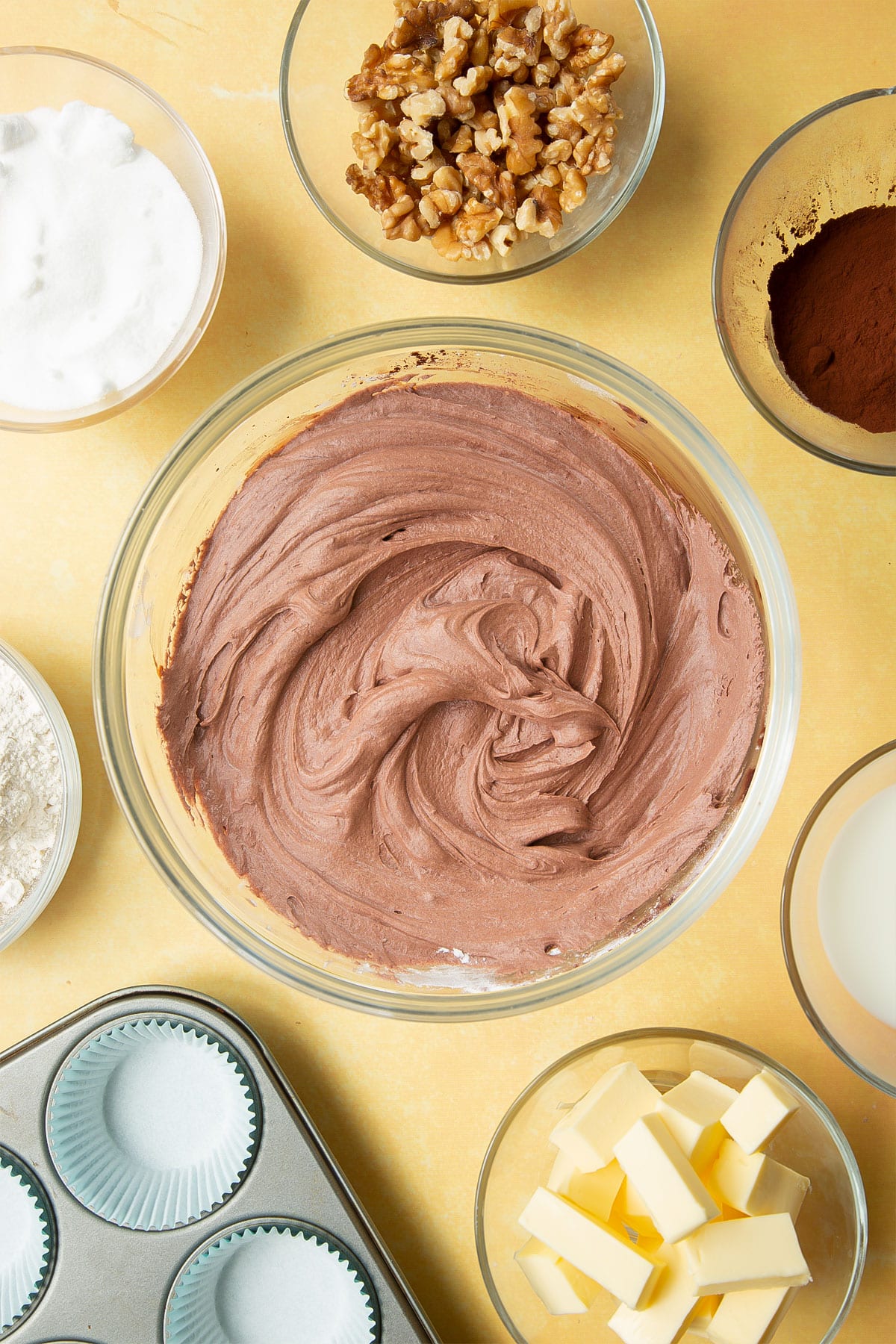 Whipped chocolate frosting in a glass mixing bowl. Ingredients for chocolate walnut cupcakes surround the bowl.