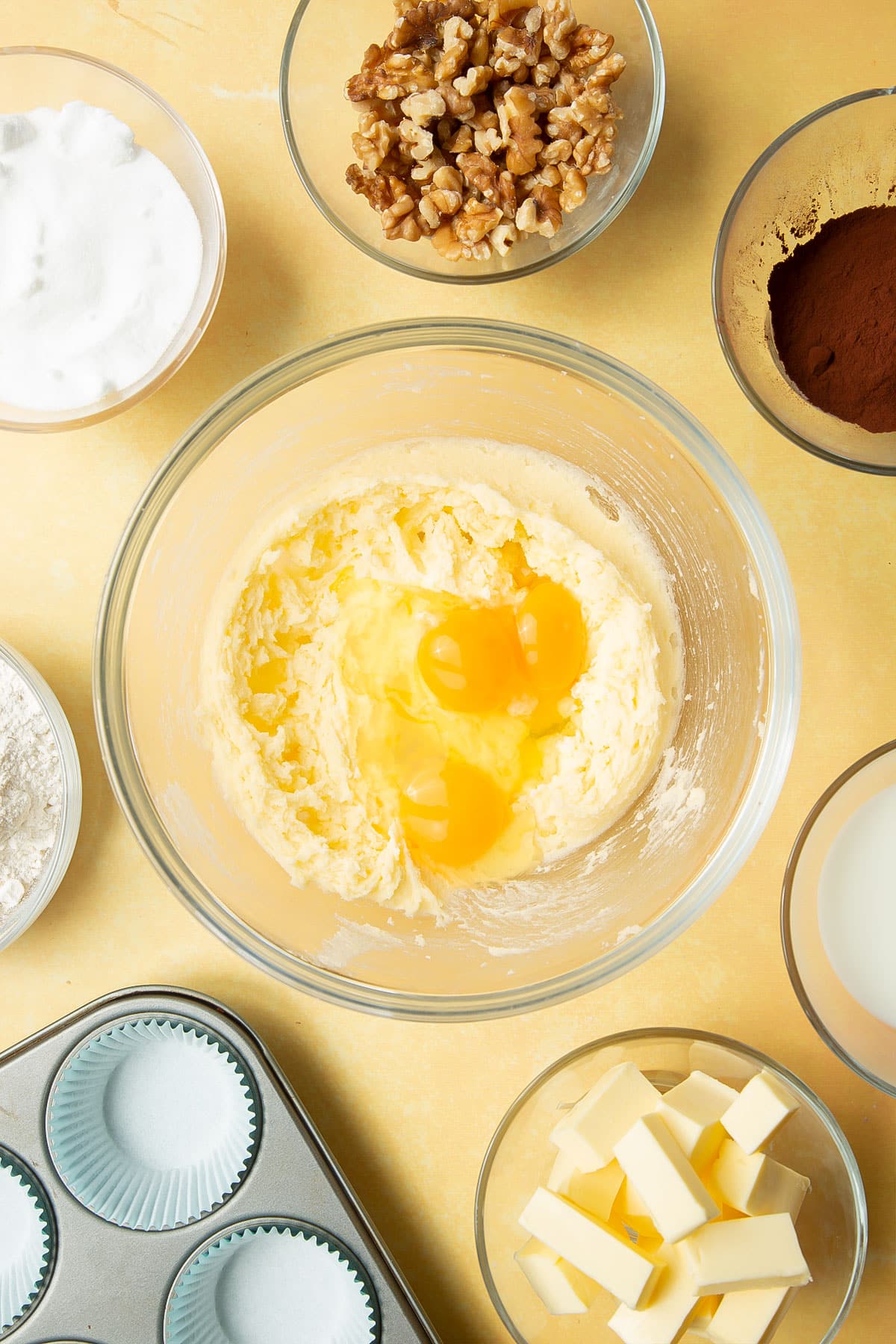 Butter and sugar, creamed together in a glass mixing bowl and topped with eggs. Ingredients for chocolate walnut cupcakes surround the bowl.