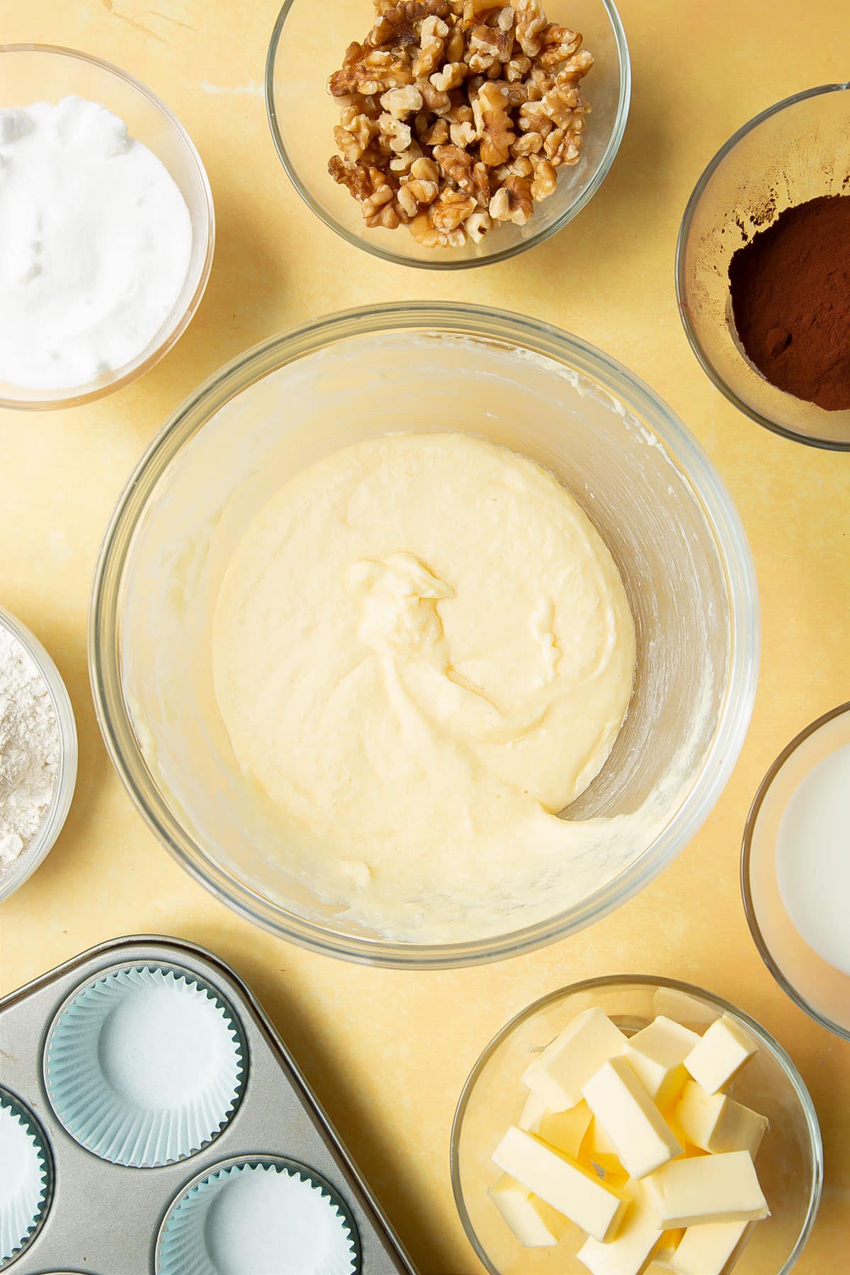 Butter, sugar and eggs beaten together in a glass mixing bowl. Ingredients for chocolate walnut cupcakes surround the bowl.