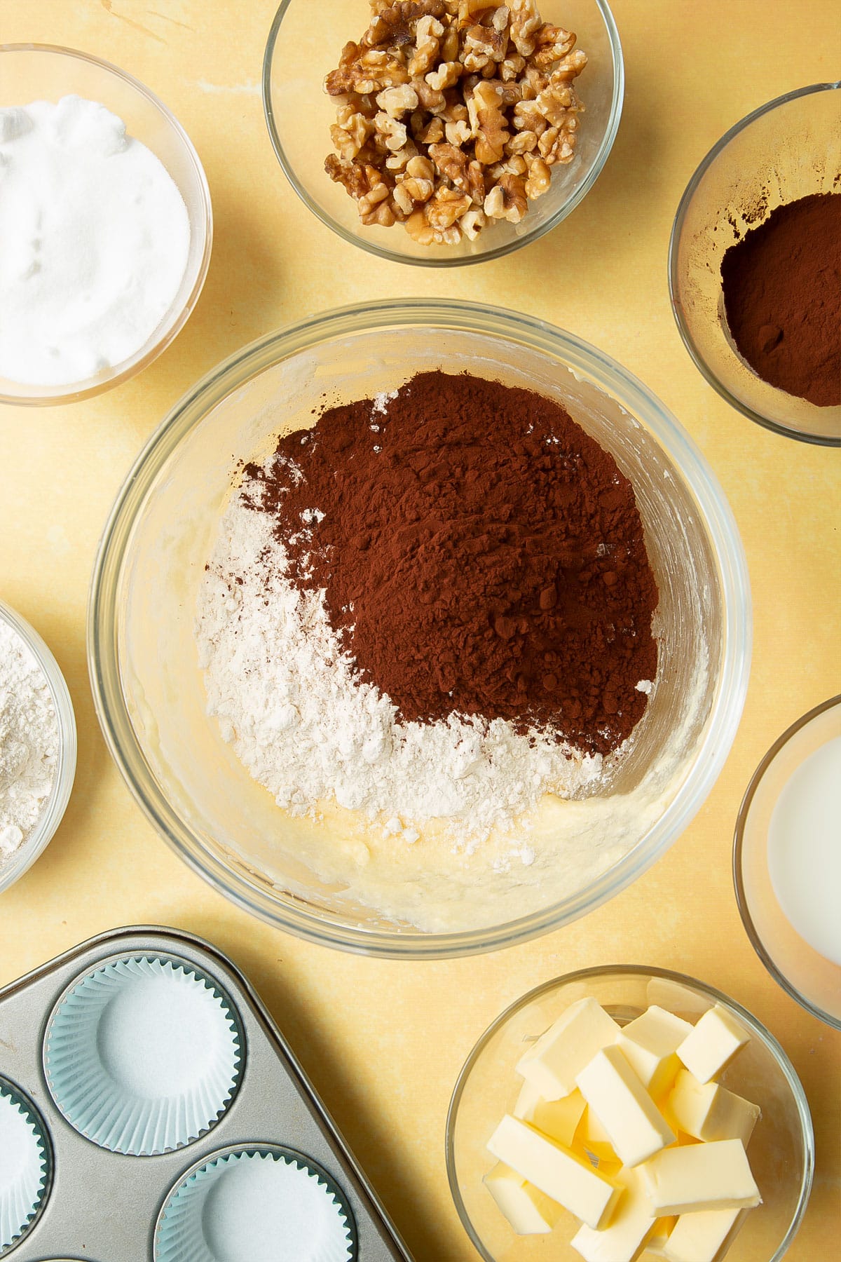 Butter, sugar and eggs creamed together in a glass mixing bowl and topped with flour and cocoa. Ingredients for chocolate walnut cupcakes surround the bowl.