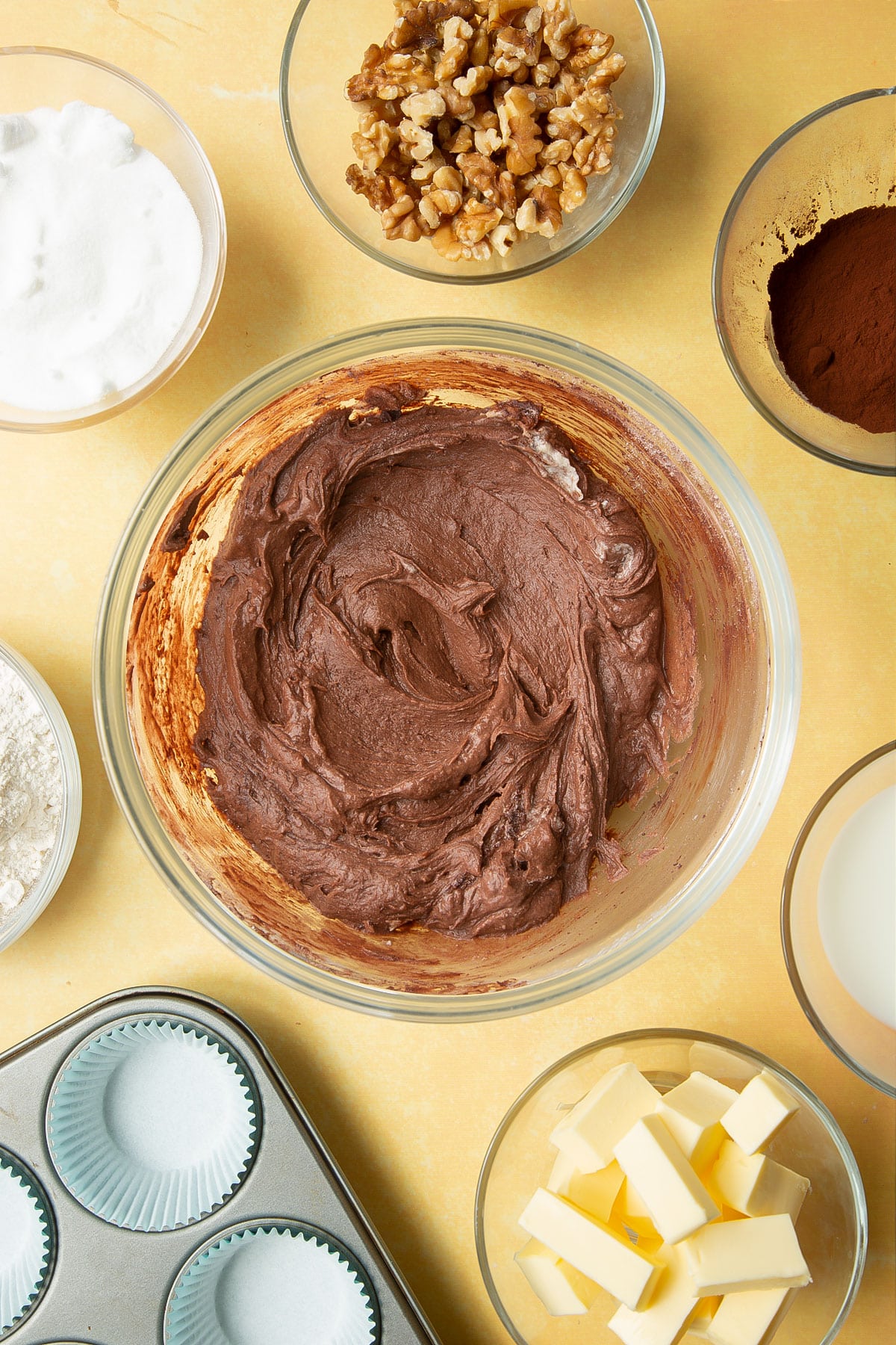 Butter, sugar, eggs, flour and cocoa mixed together in a glass mixing bowl. Ingredients for chocolate walnut cupcakes surround the bowl.