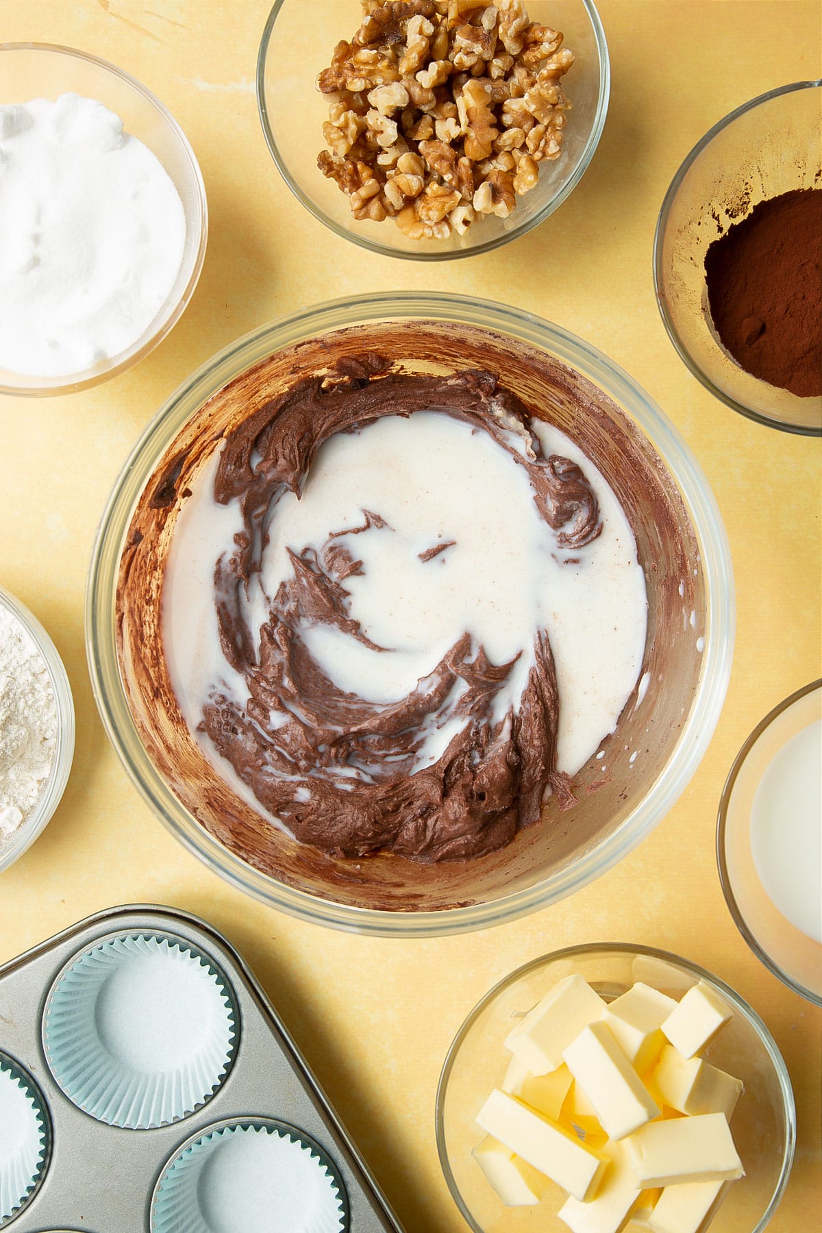 Butter, sugar, eggs, flour and cocoa mixed together in a glass mixing bowl with milk on top. Ingredients for chocolate walnut cupcakes surround the bowl.