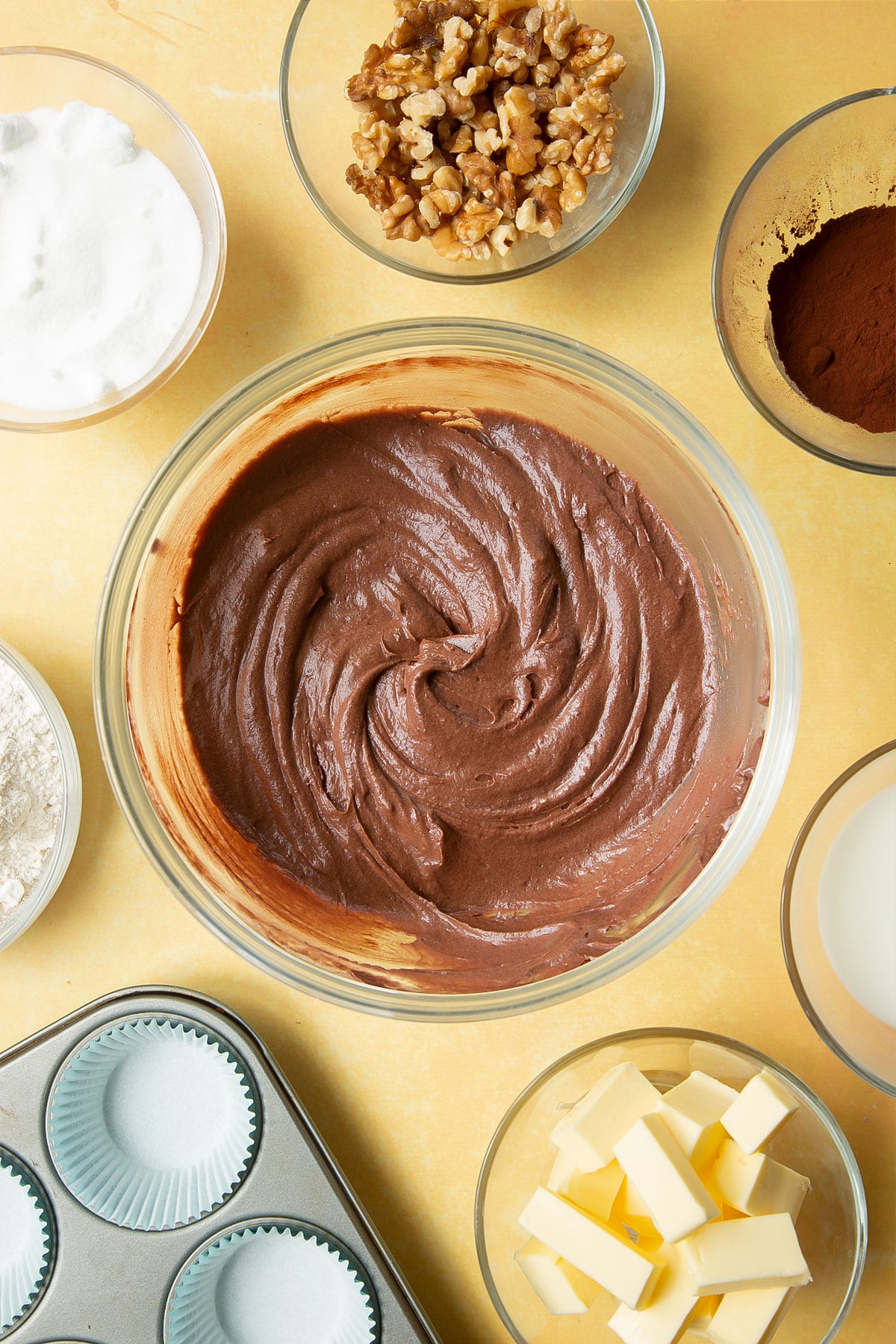 Chocolate cake batter in a glass mixing bowl. Ingredients for chocolate walnut cupcakes surround the bowl.