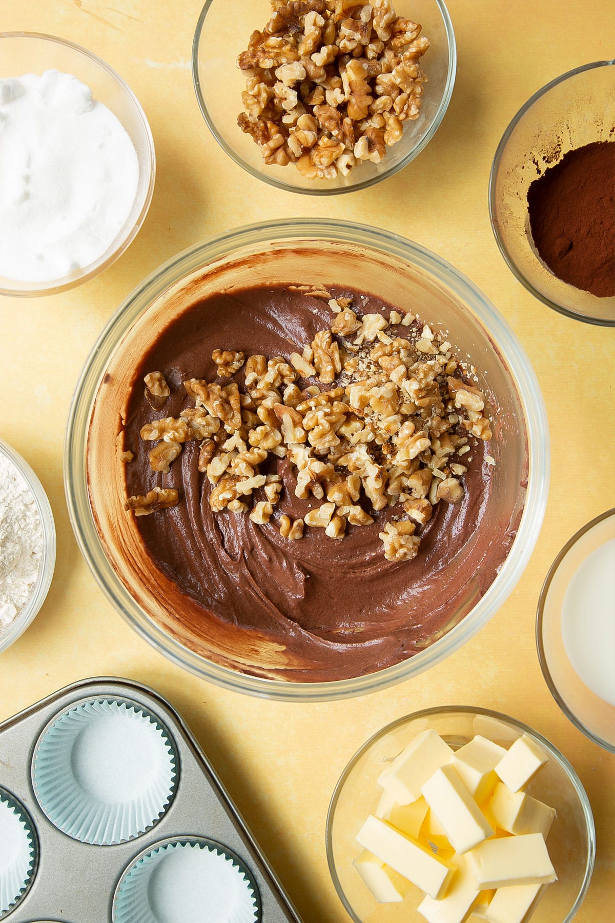 Chocolate cupcake batter in a glass mixing bowl with chopped walnuts on top. Ingredients for chocolate walnut cupcakes surround the bowl.