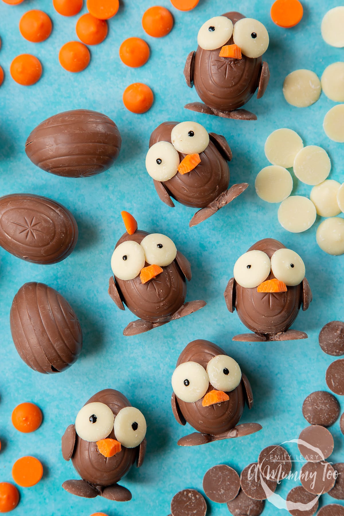 Five chocolate chicks made from creme eggs and chocolate buttons, laying on a blue background, surrounded by milk chocolate buttons, white chocolate buttons and chocolate orange buttons. 
