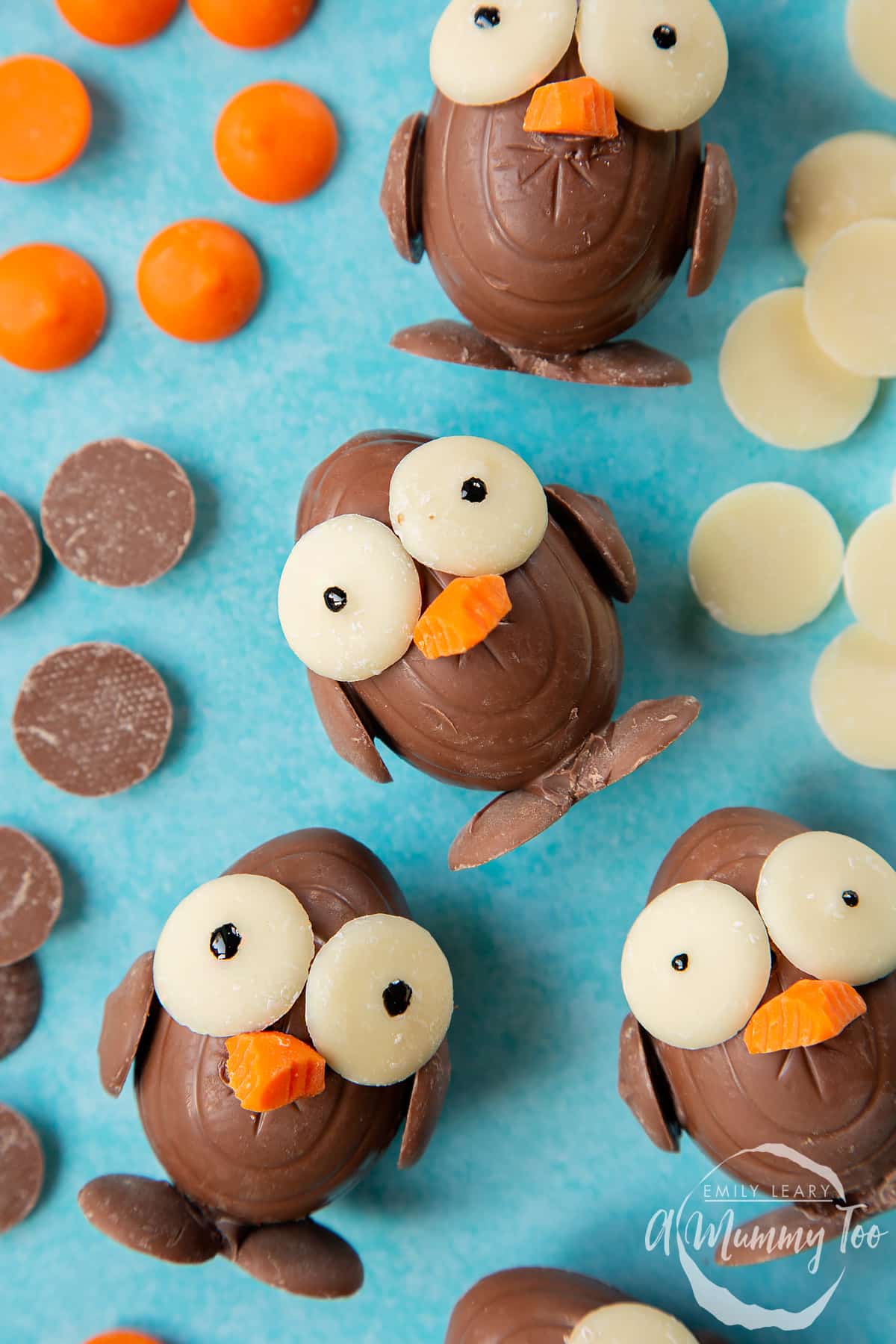 Chocolate chicks made from creme eggs and chocolate buttons, laying on a blue background, surrounded by milk chocolate buttons, white chocolate buttons and chocolate orange buttons. 