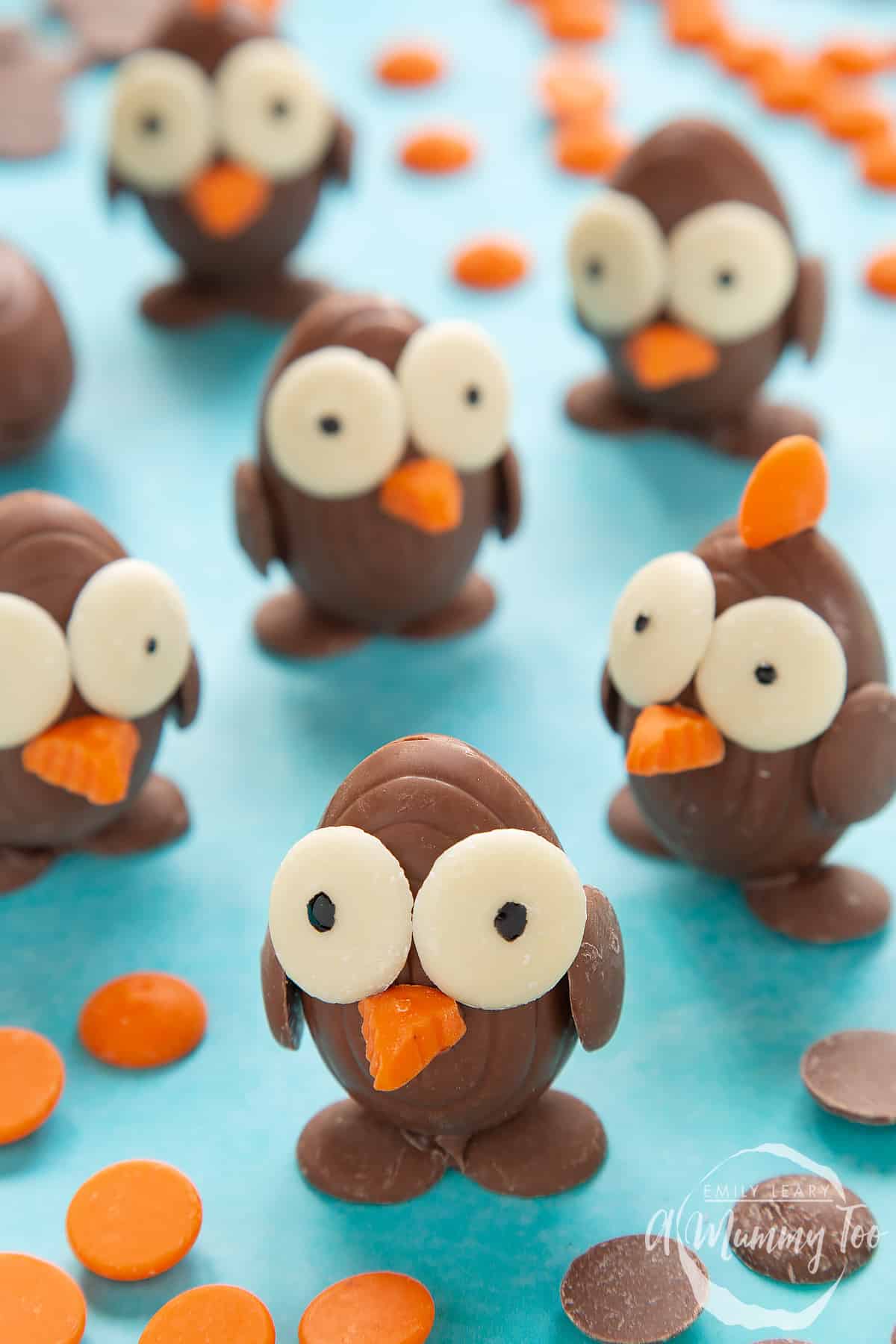 Chocolate chicks made from creme eggs and chocolate buttons, standing on a blue background, surrounded by milk chocolate buttons, white chocolate buttons and chocolate orange buttons. One of the chicks in the background has an orange crest. 