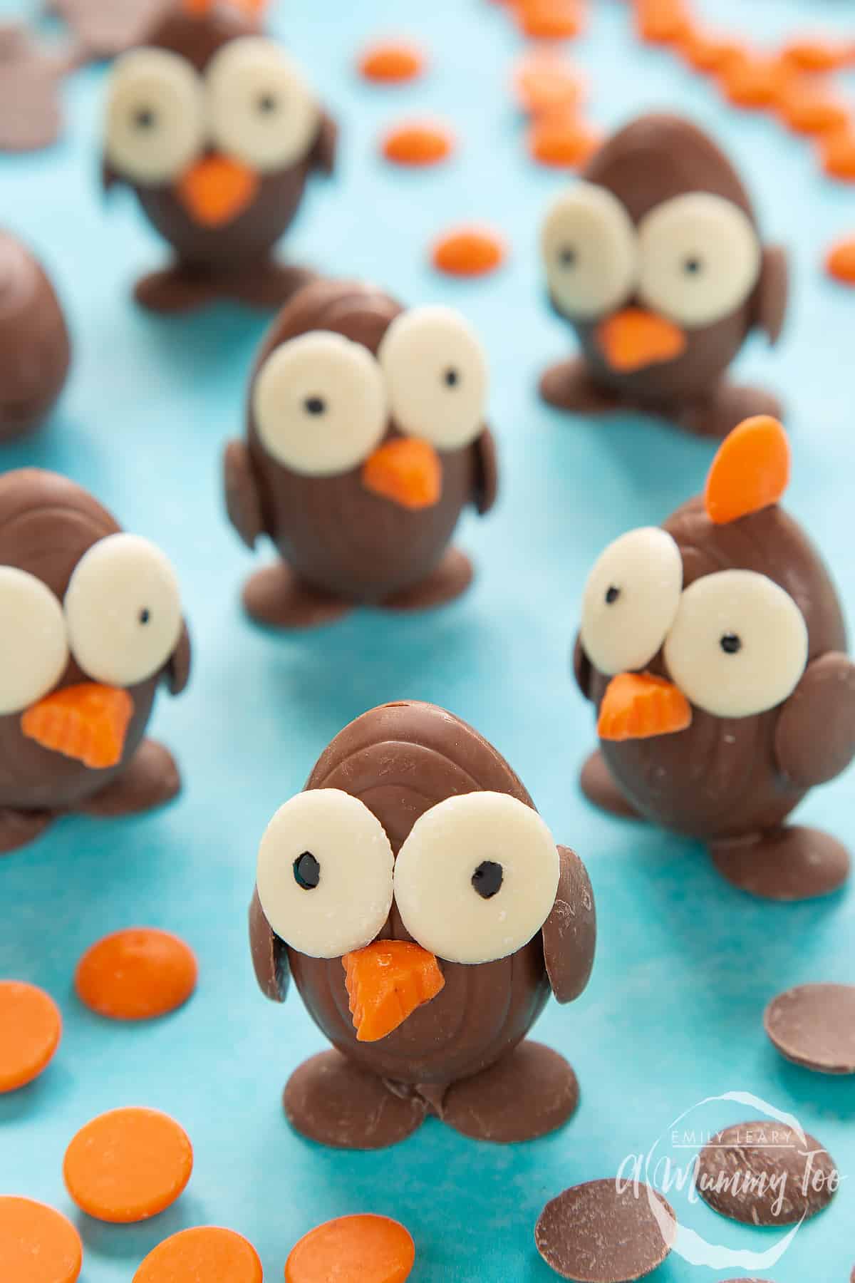 Chocolate chicks made from creme eggs and chocolate buttons, standing on a blue background, surrounded by milk chocolate buttons, white chocolate buttons and chocolate orange buttons. One of the chicks in the background has an orange crest.