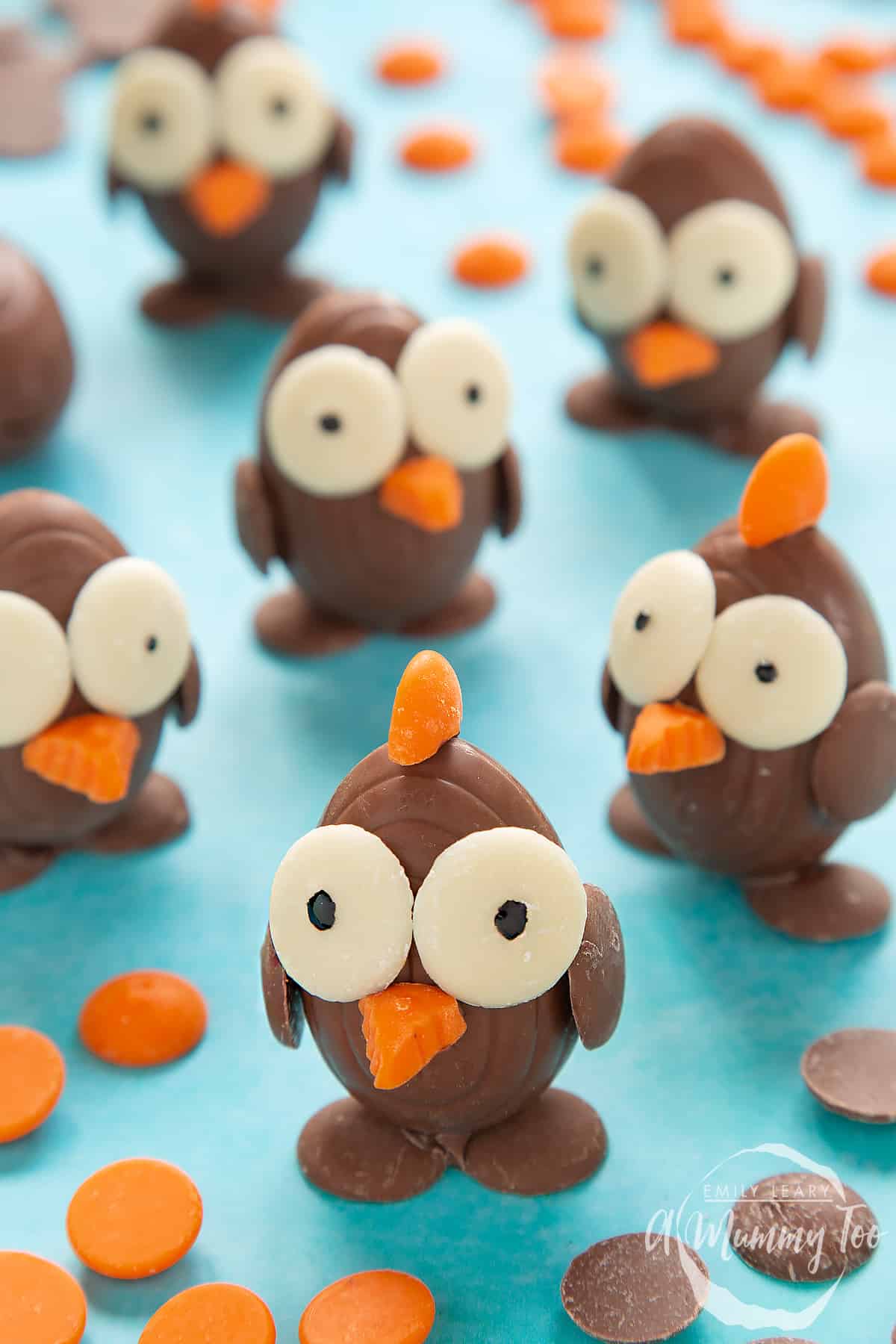 Chocolate chicks made from creme eggs and chocolate buttons, standing on a blue background, surrounded by milk chocolate buttons, white chocolate buttons and chocolate orange buttons. 