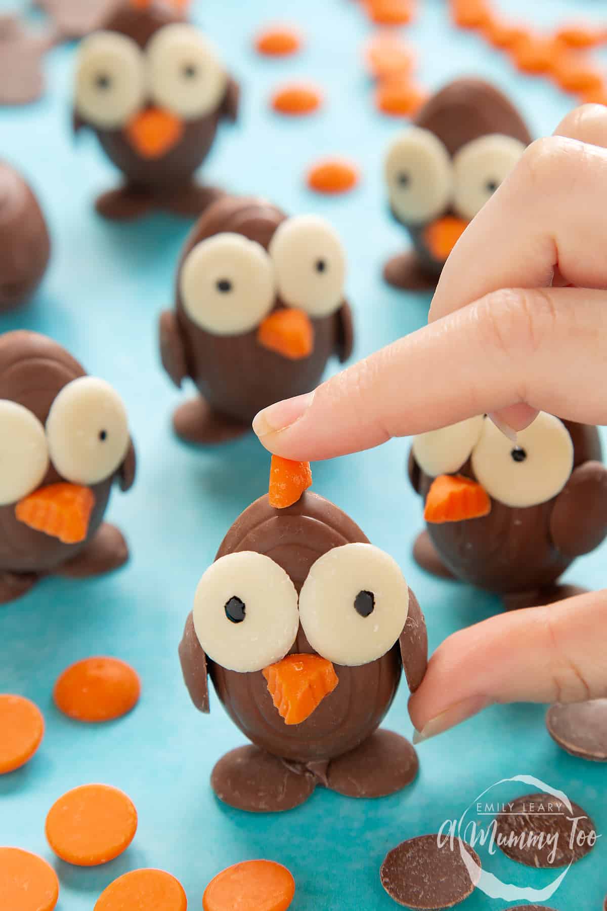 Chocolate chicks made from creme eggs and chocolate buttons, standing on a blue background, surrounded by milk chocolate buttons, white chocolate buttons and chocolate orange buttons. A hand is holding the foremost chick.