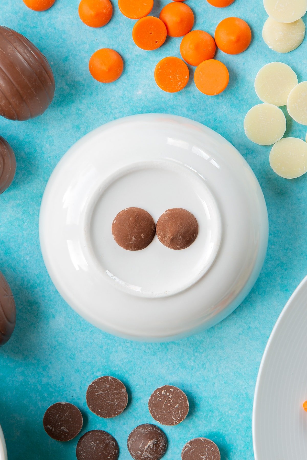 Two chocolate buttons sit on an upturned white bowl, surrounded by ingredients to make chocolate chicks.