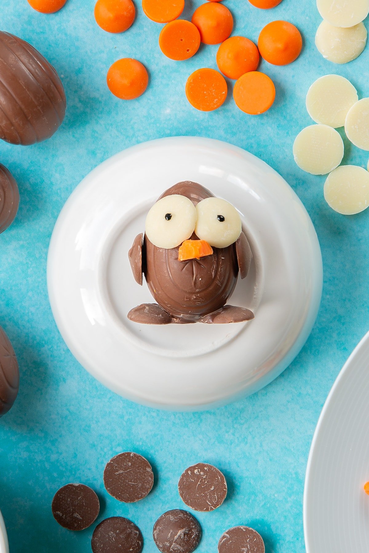 A creme egg is decorated with chocolate buttons to resemble an Easter chick. Pupils are drawn onto the eyes with black icing. The chick rests on a bowl, surrounded by ingredients to make chocolate chicks.