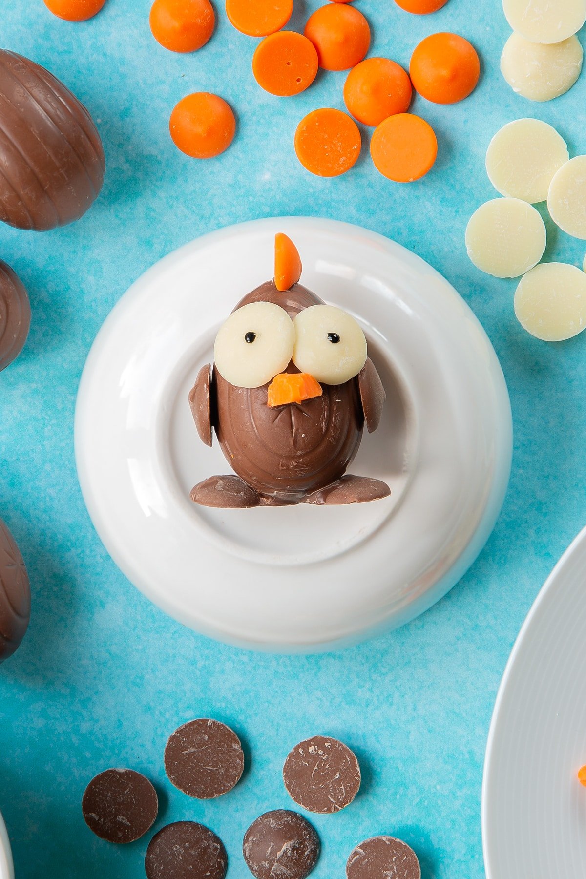 A creme egg is decorated with chocolate buttons to resemble an Easter chick. Half an orange button is stuck to the top to resemble a crest. The chick rests on a bowl, surrounded by ingredients to make chocolate chicks.