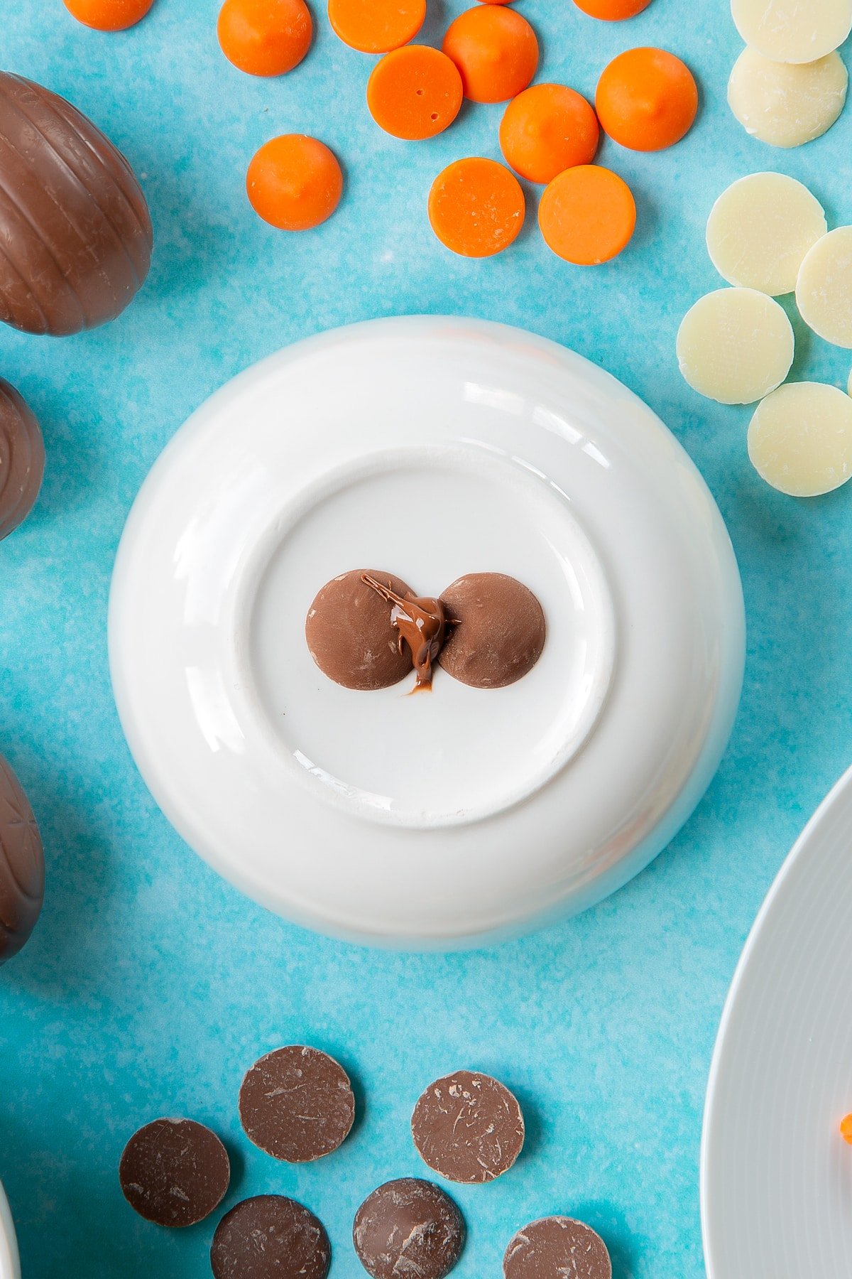 Two chocolate buttons topped with a little melted chocolate sit on an upturned white bowl, surrounded by ingredients to make chocolate chicks.