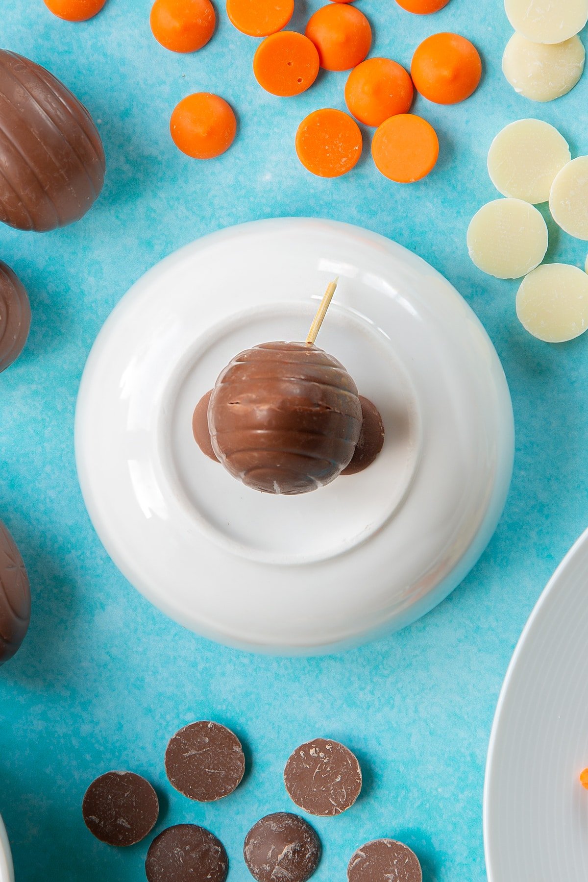 Two chocolate buttons topped with a creme egg sit on an upturned white bowl, surrounded by ingredients to make chocolate chicks.