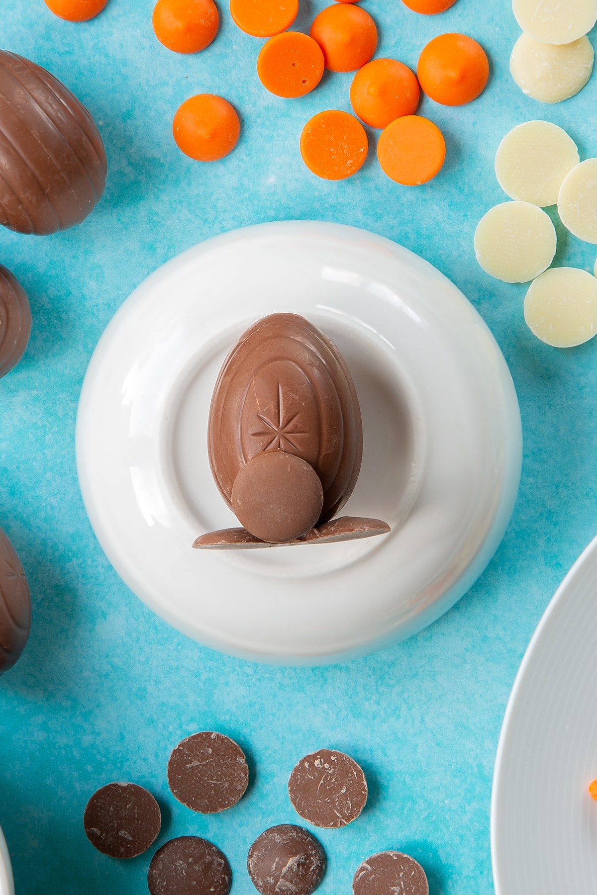 Two chocolate buttons stuck to the bottom of a creme egg to resemble feet. The egg lays on an upturned white bowl. Another chocolate button is stuck to the egg to resemble a tail. The bowl is surrounded by ingredients to make chocolate chicks.