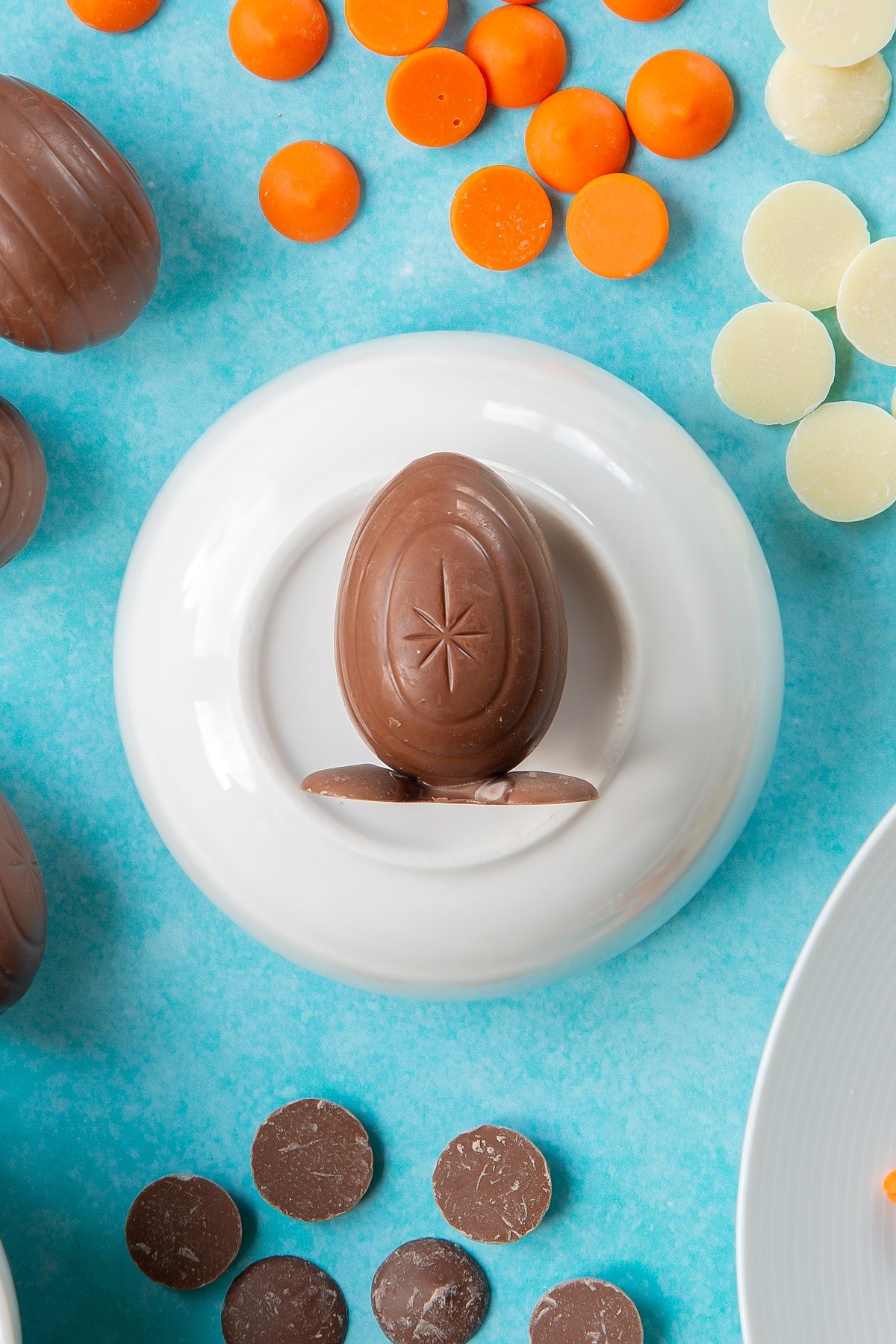 Two chocolate buttons stuck to the bottom of a creme egg to resemble feet. The egg lays on an upturned white bowl, surrounded by ingredients to make chocolate chicks.