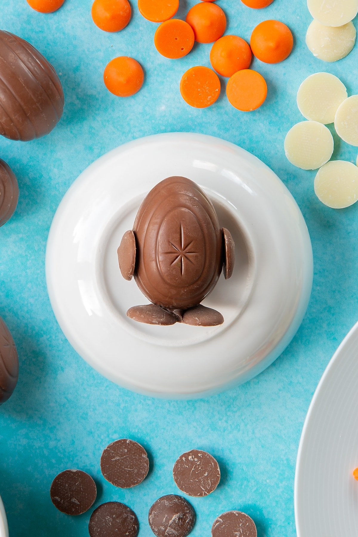 Two chocolate buttons stuck to the bottom of a creme egg to resemble feet. The egg lays on an upturned white bowl. Two more chocolate buttons are stuck to the sides of the egg to resemble wings. The bowl is surrounded by ingredients to make chocolate chicks.