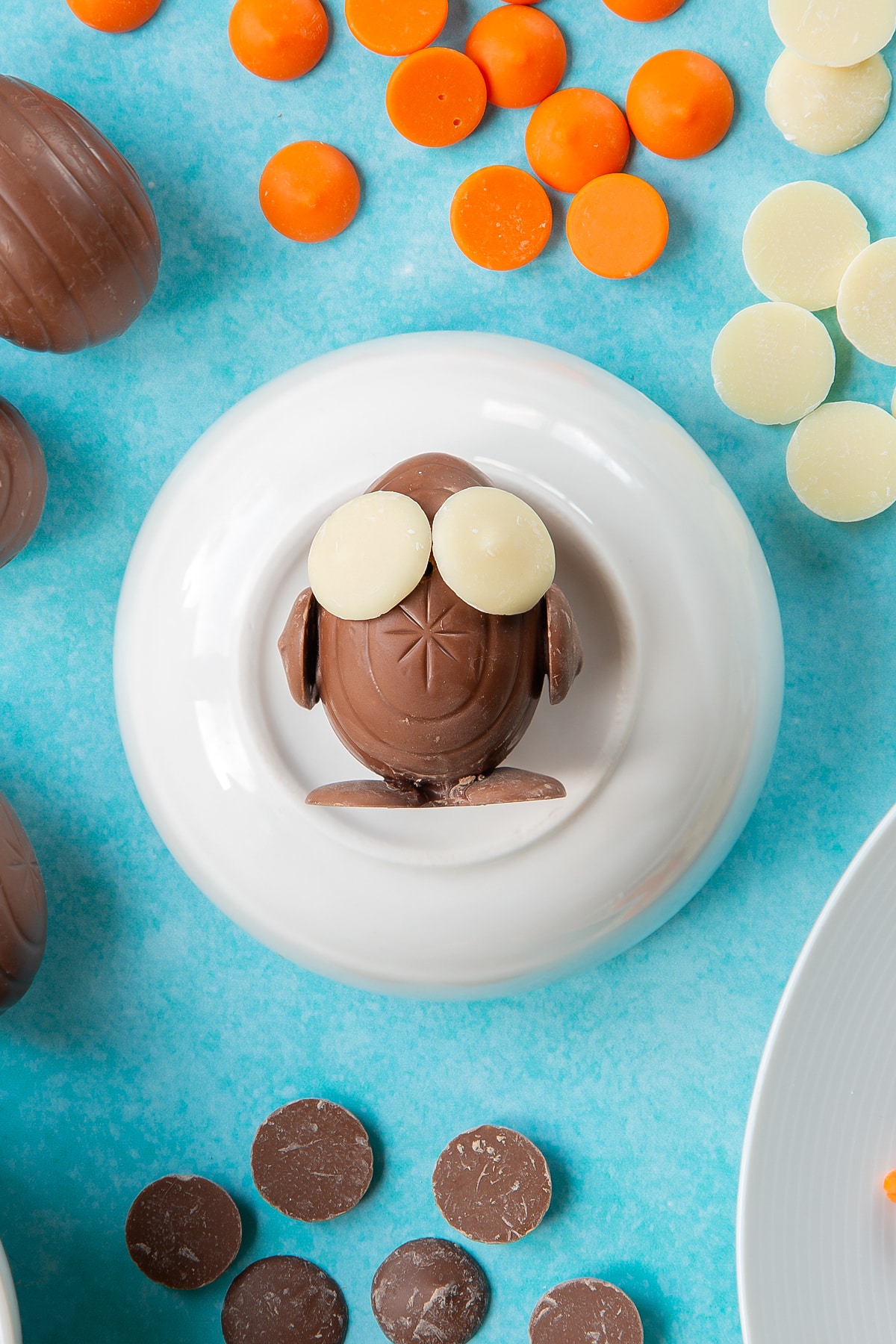 Two chocolate buttons stuck to the bottom of a creme egg to resemble feet. The egg lays on an upturned white bowl. Two more chocolate buttons are stuck to the sides of the egg to resemble wings, while two white chocolate buttons form eyes. The bowl is surrounded by ingredients to make chocolate chicks.