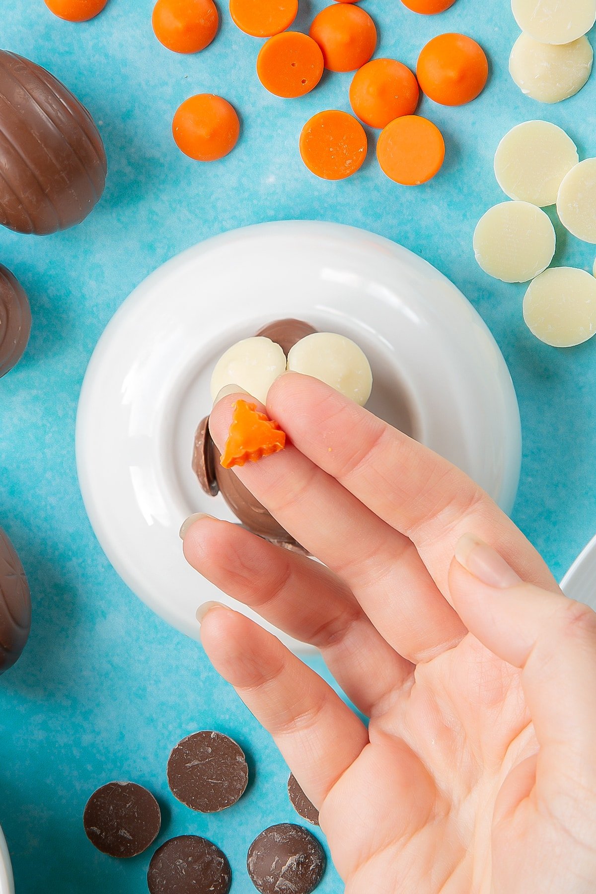 A hand holds a small chocolate orange triangle. Below, a creme egg lays on an upturned white bowl. The bowl is surrounded by ingredients to make chocolate chicks.