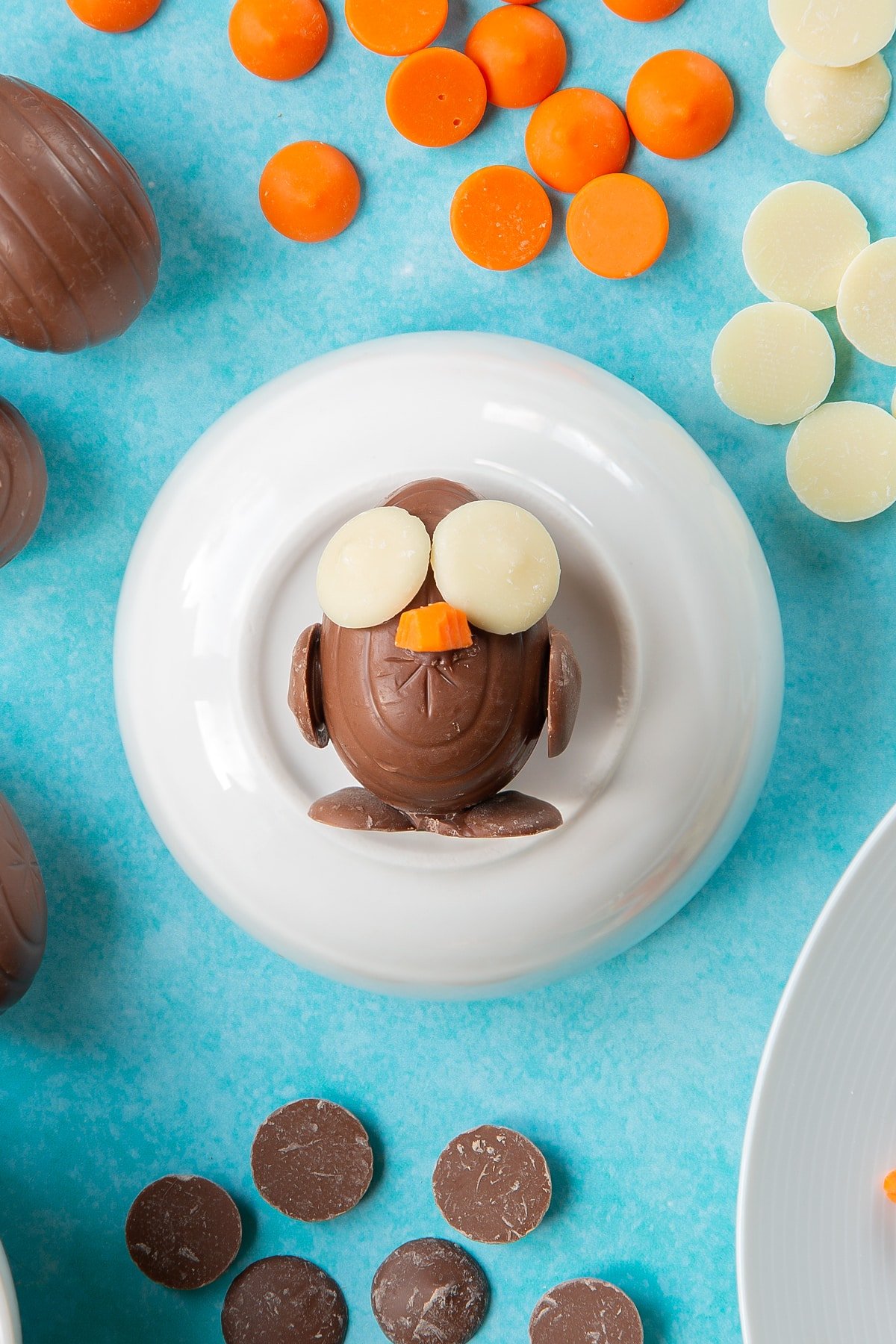 A creme egg is decorated with chocolate buttons to resemble an Easter chick. It rests on a bowl, surrounded by ingredients to make chocolate chicks.