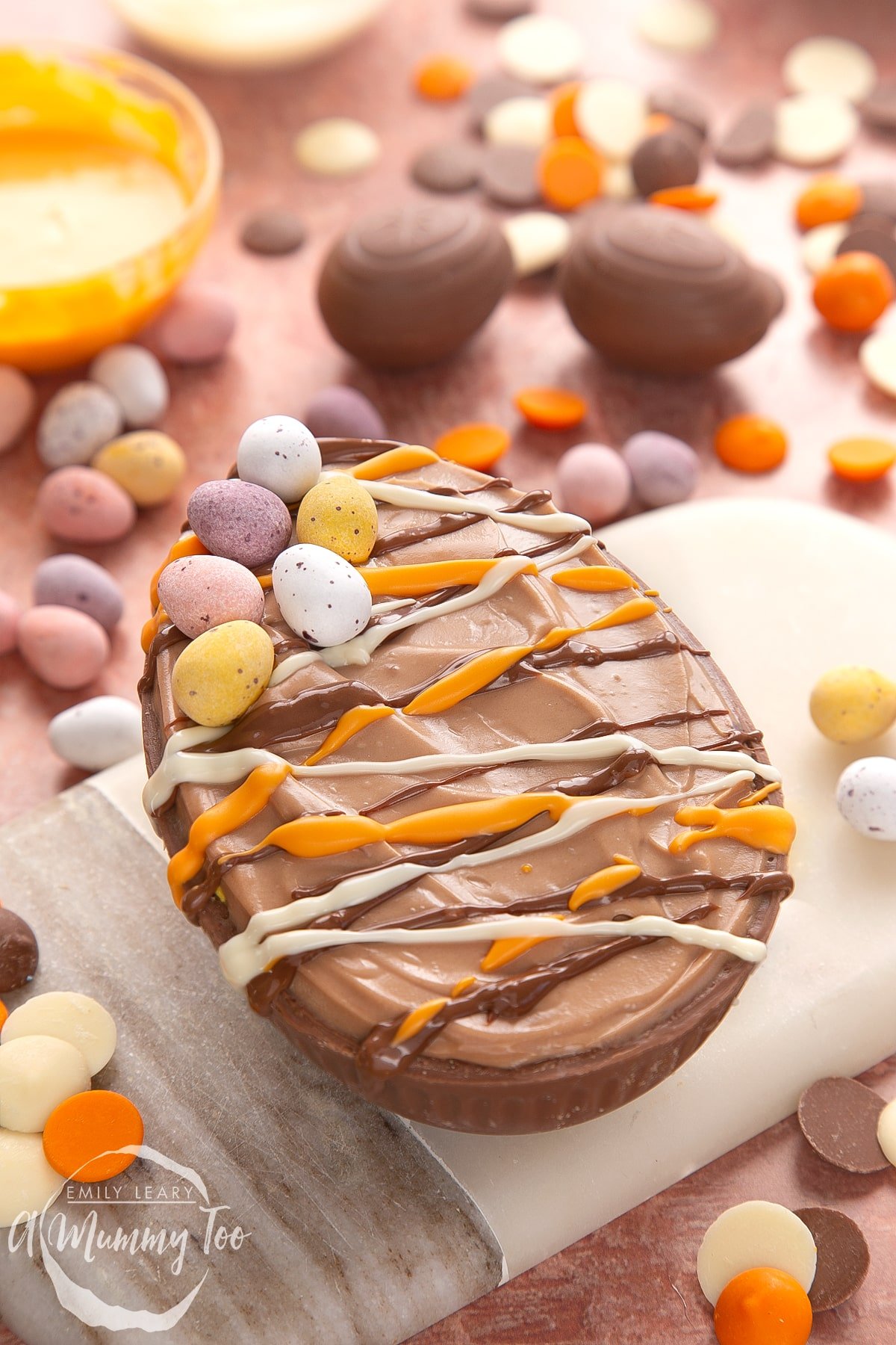 An Easter egg cheesecake on a small marble board. The chocolate cheesecake filling sits in a chocolate Easter egg shell, drizzled with white, milk and orange chocolate.