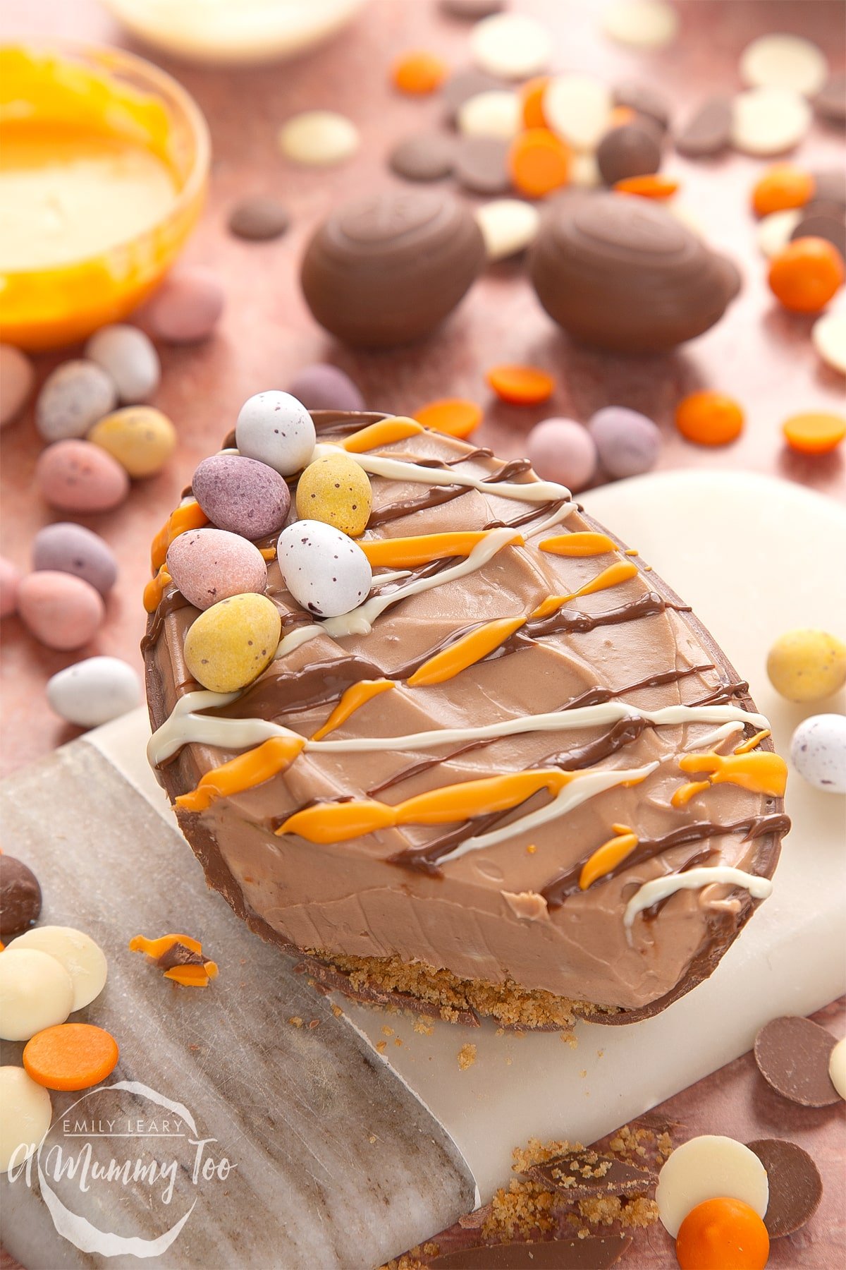 An Easter egg cheesecake on a small marble board. The chocolate cheesecake filling sits in a chocolate Easter egg shell, drizzled with white, milk and orange chocolate. The egg has been cut to show the hidden biscuit base.
