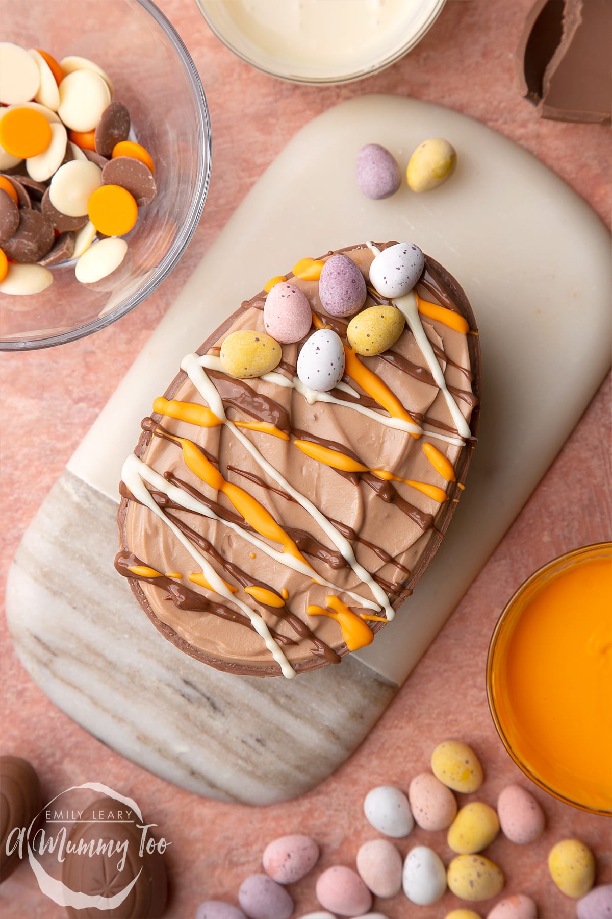 An Easter egg cheesecake on a small marble board, seen from above. The chocolate cheesecake filling sits in a chocolate Easter egg shell, drizzled with white, milk and orange chocolate.