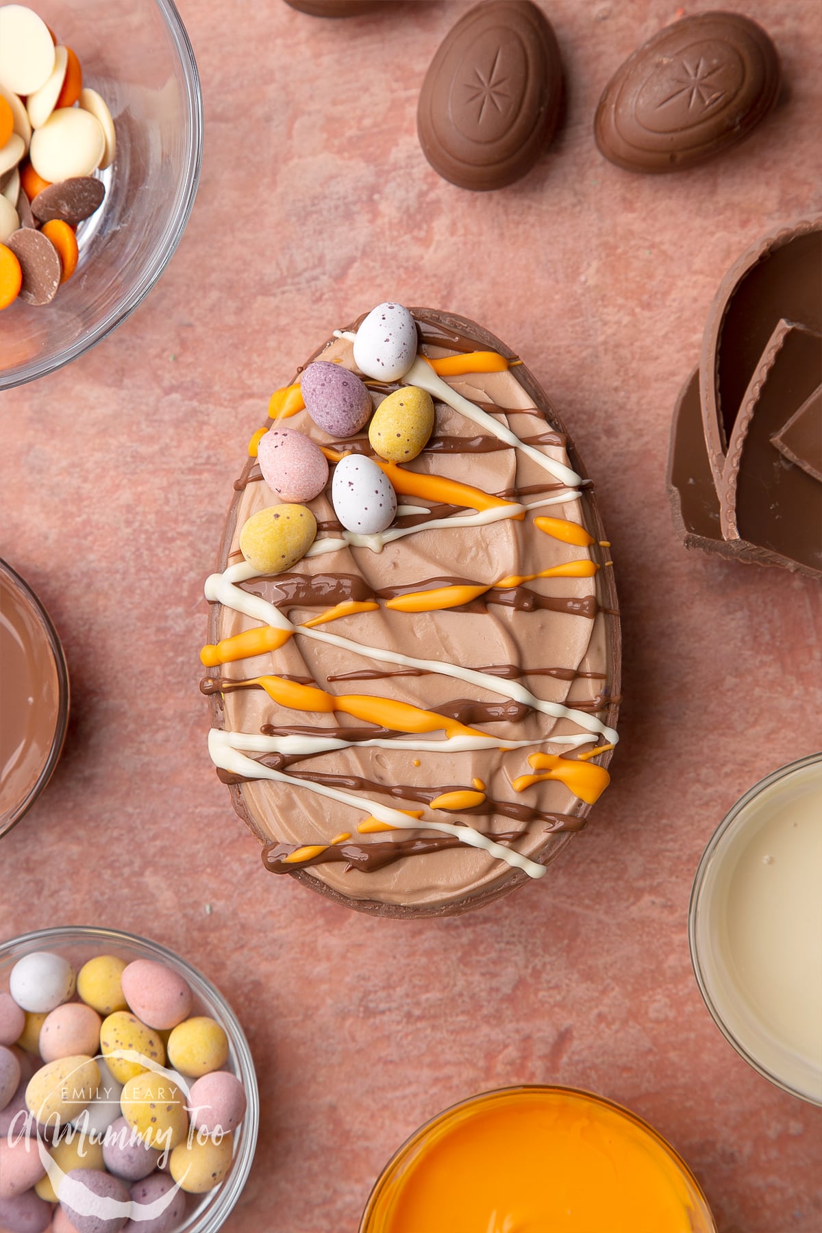 An Easter egg cheesecake on a small marble board. The chocolate cheesecake filling sits in a chocolate Easter egg shell, drizzled with white, milk and orange chocolate. The egg is surrounded by ingredient such as mini eggs.