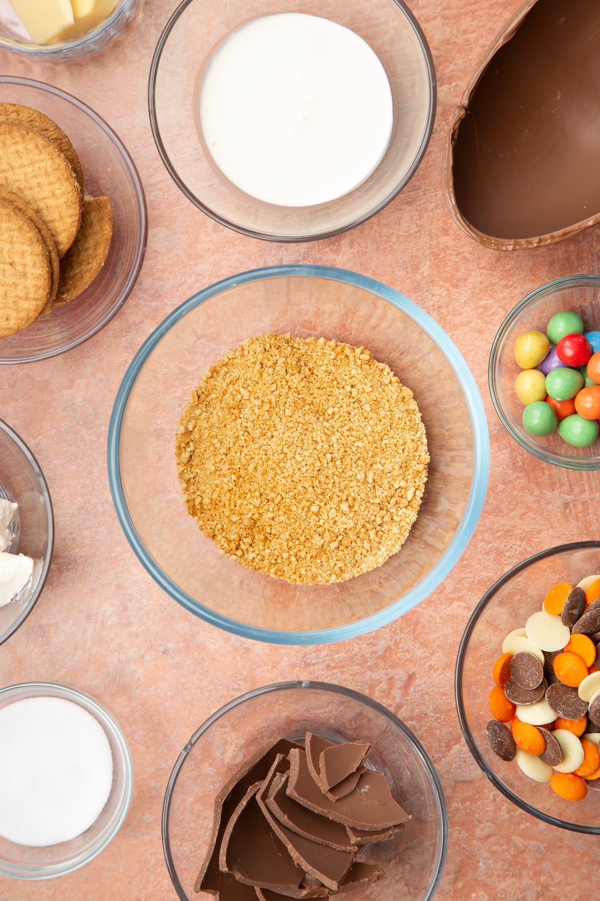 A glass bowl containing crushed digestive biscuits. The bowl is surrounded by ingredients to make Easter Egg cheesecake.