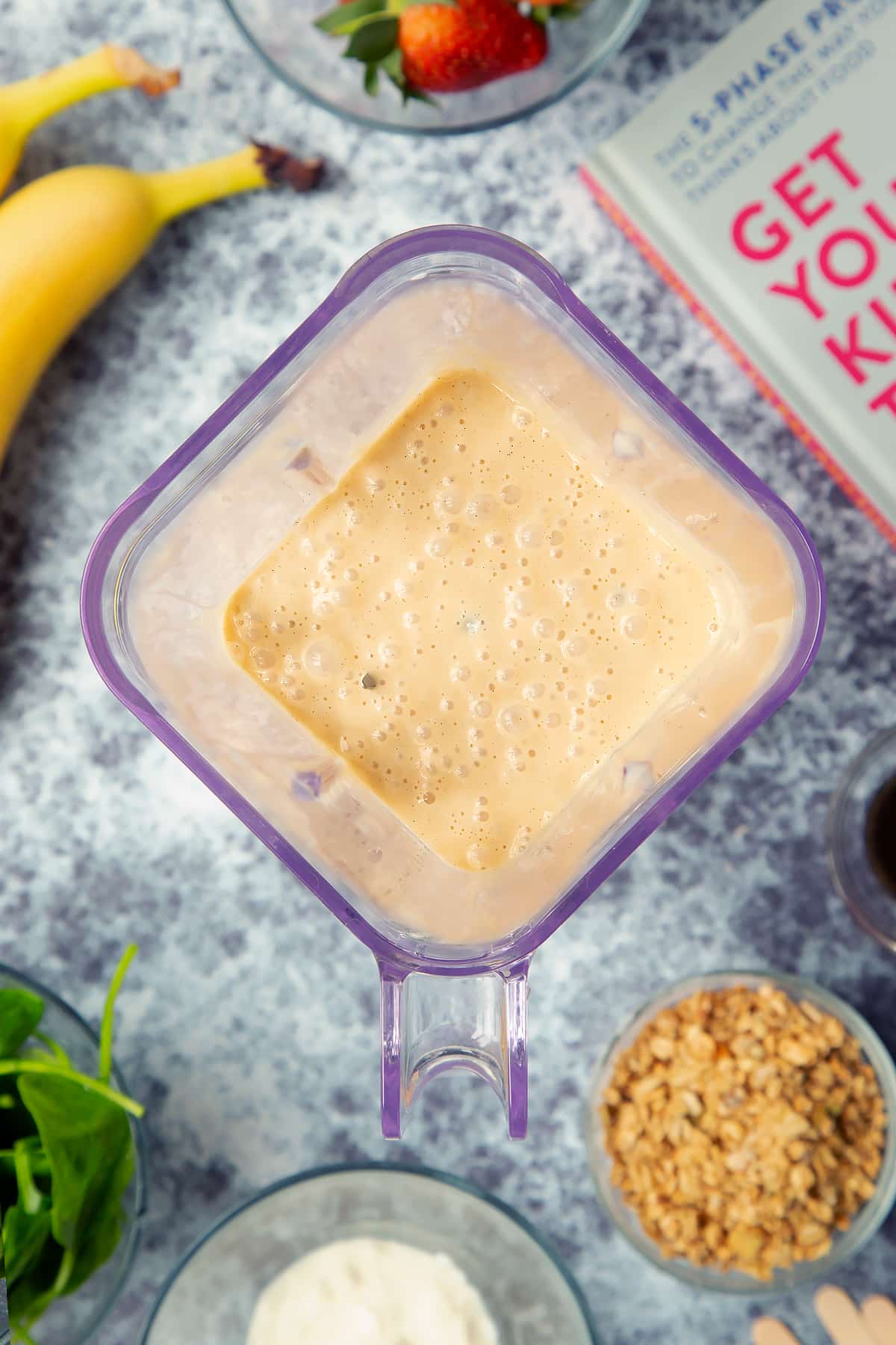 A blender is shown from above, containing yogurt, vanilla and bananas, blended together. The blender is surrounded by ingredients for fruit and veg lollies.