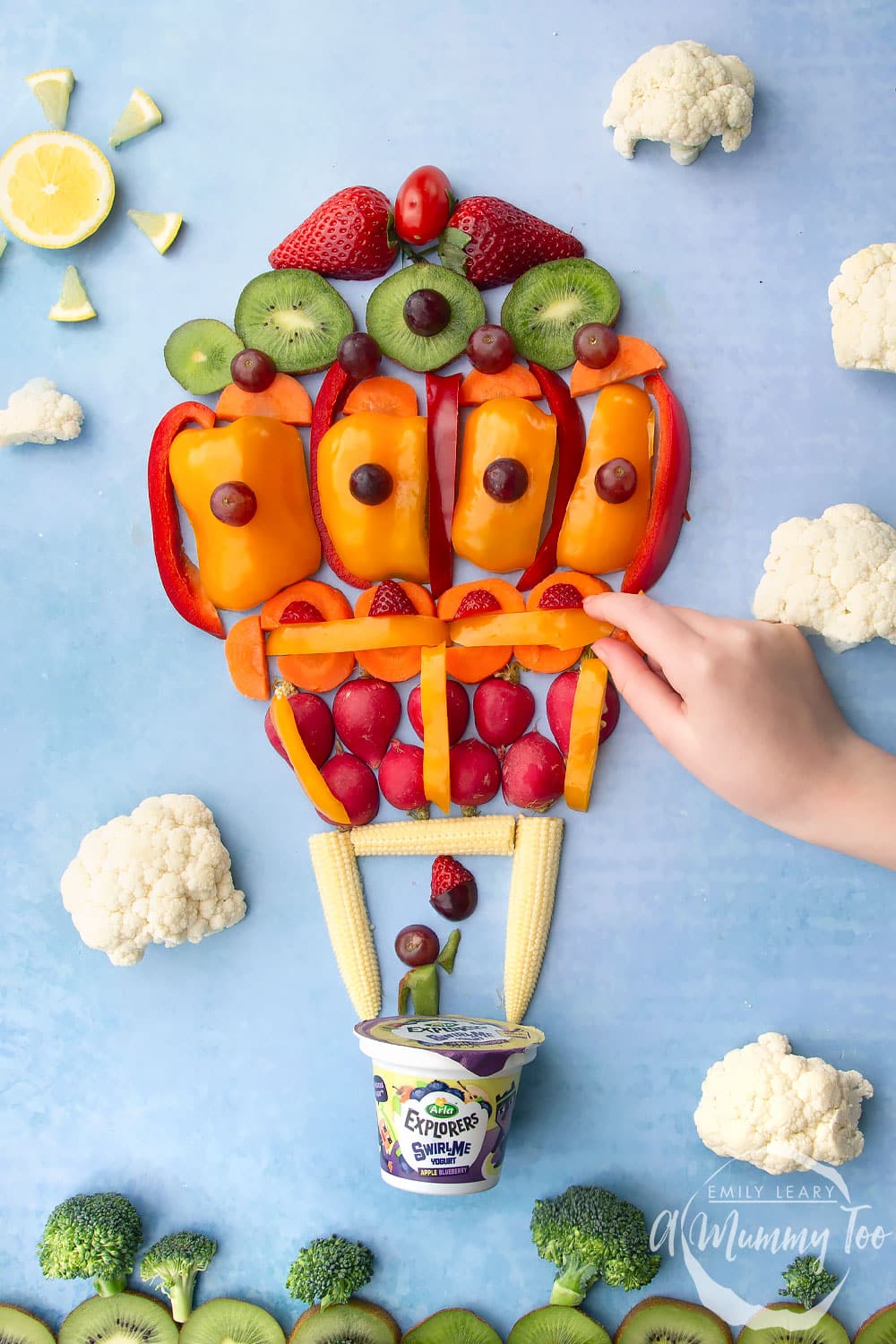 Overhead shot of a flatlay of fruit and vegetables in the shape of a hot air balloon flying in the sky. A hand is reaching in from the right hand side to grab a slice of pepper.