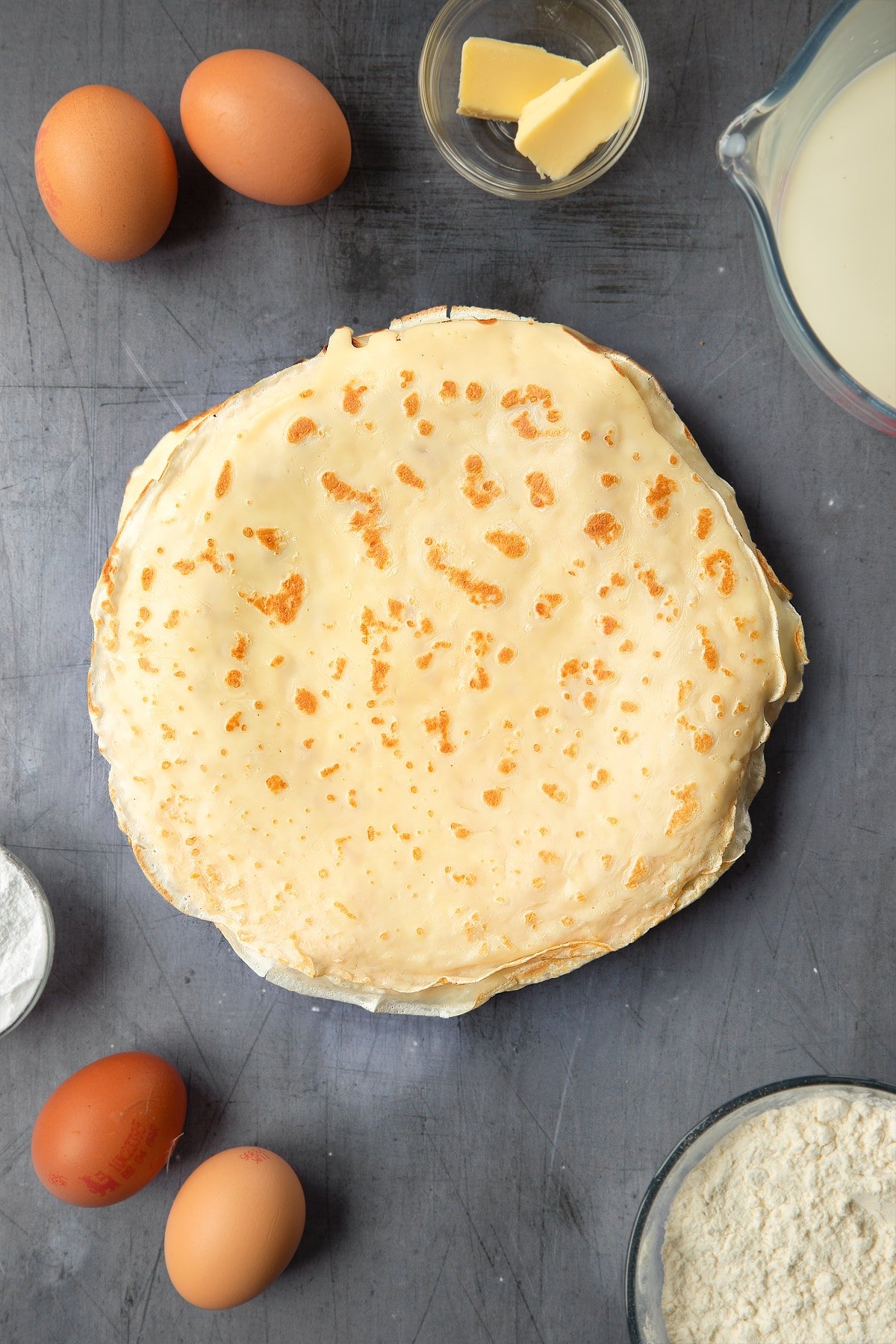 A stacked of freshly cooked crepes on a plate. The plate is surrounded by ingredients to make fun pancake ideas.