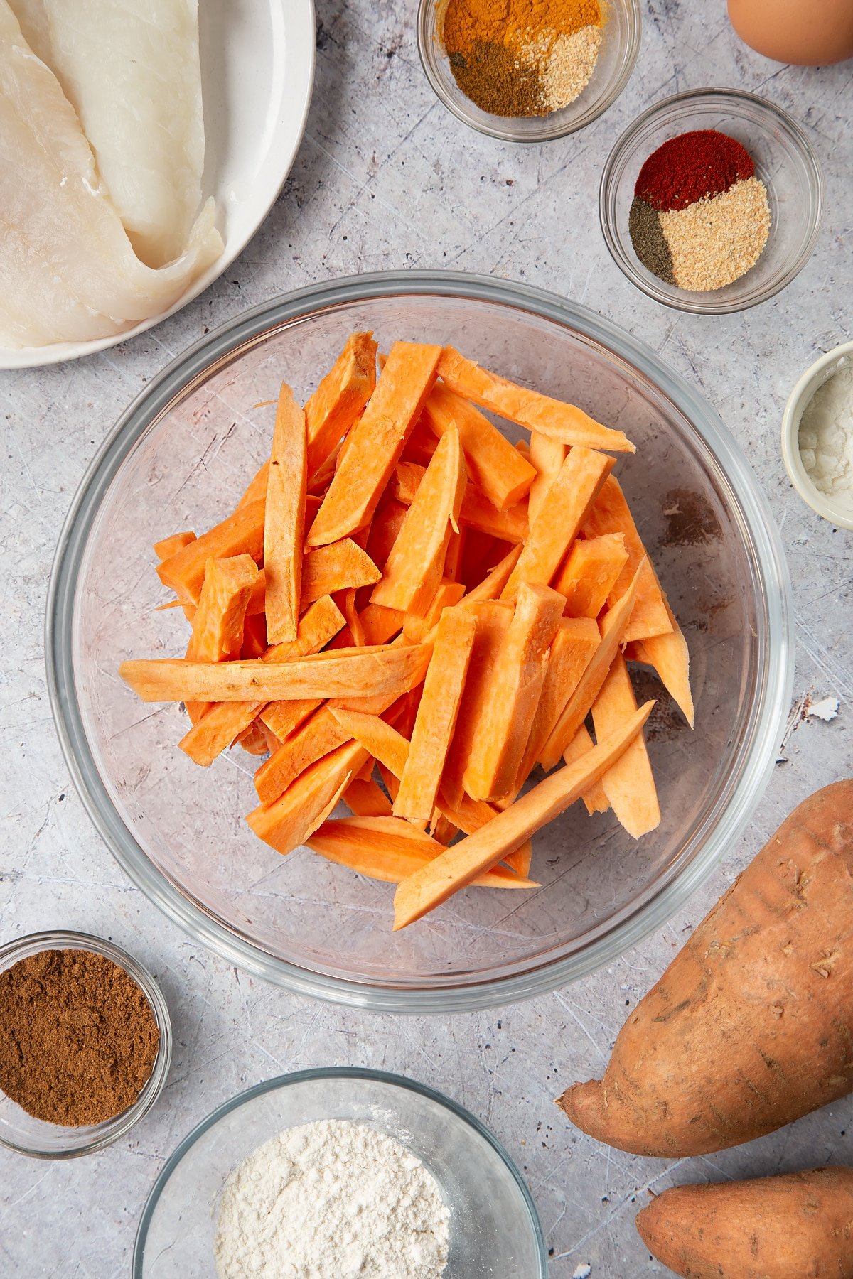 Sweet potato cut into fries and placed in a glass bowl. Surrounding the bowl is ingredients for sweet potato chips.