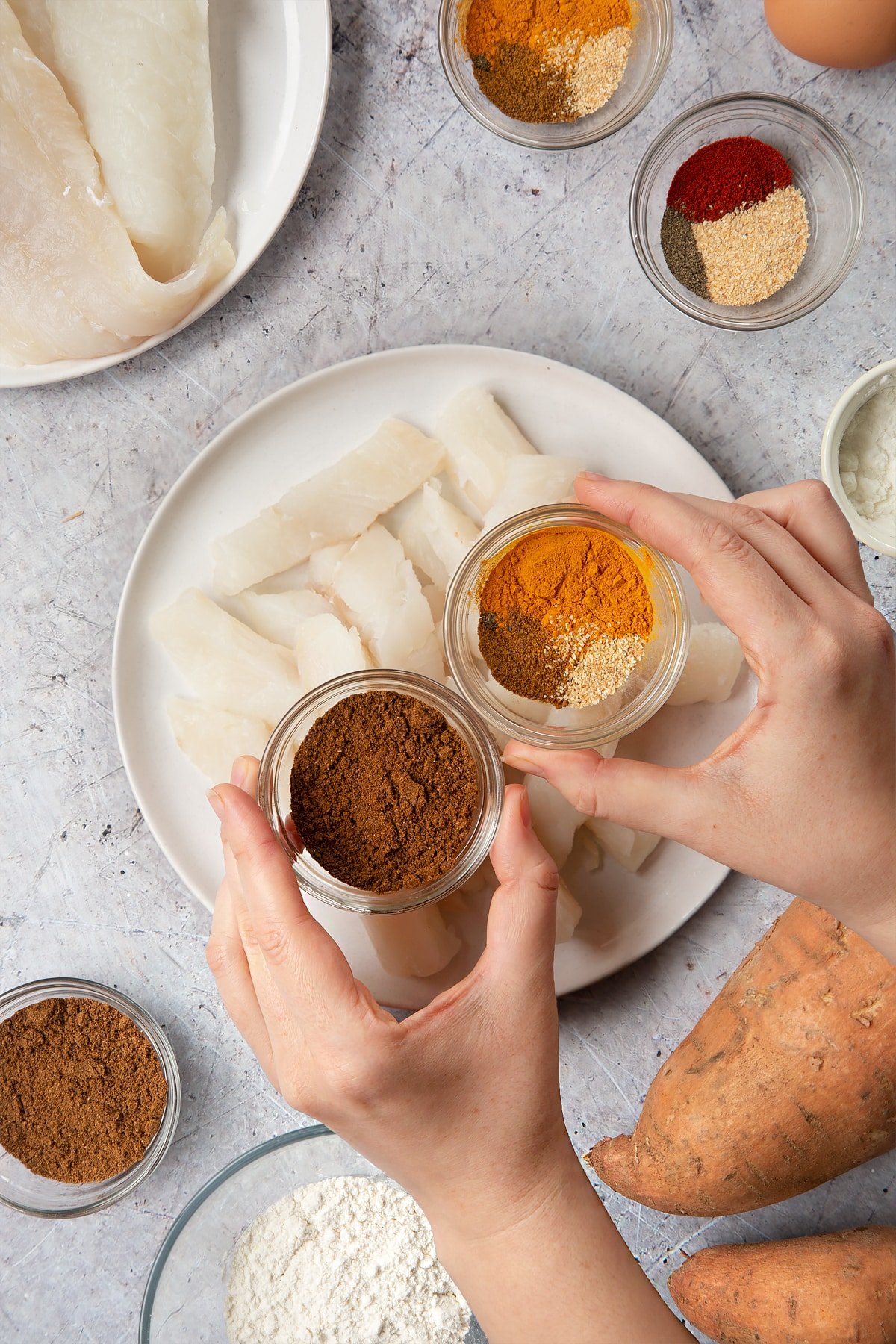 Cod fillets on a white plate, cut into fingers. Above, two hands hold two small bowls containing curry spices. Surrounding the plate is ingredients for spicy fish sticks.