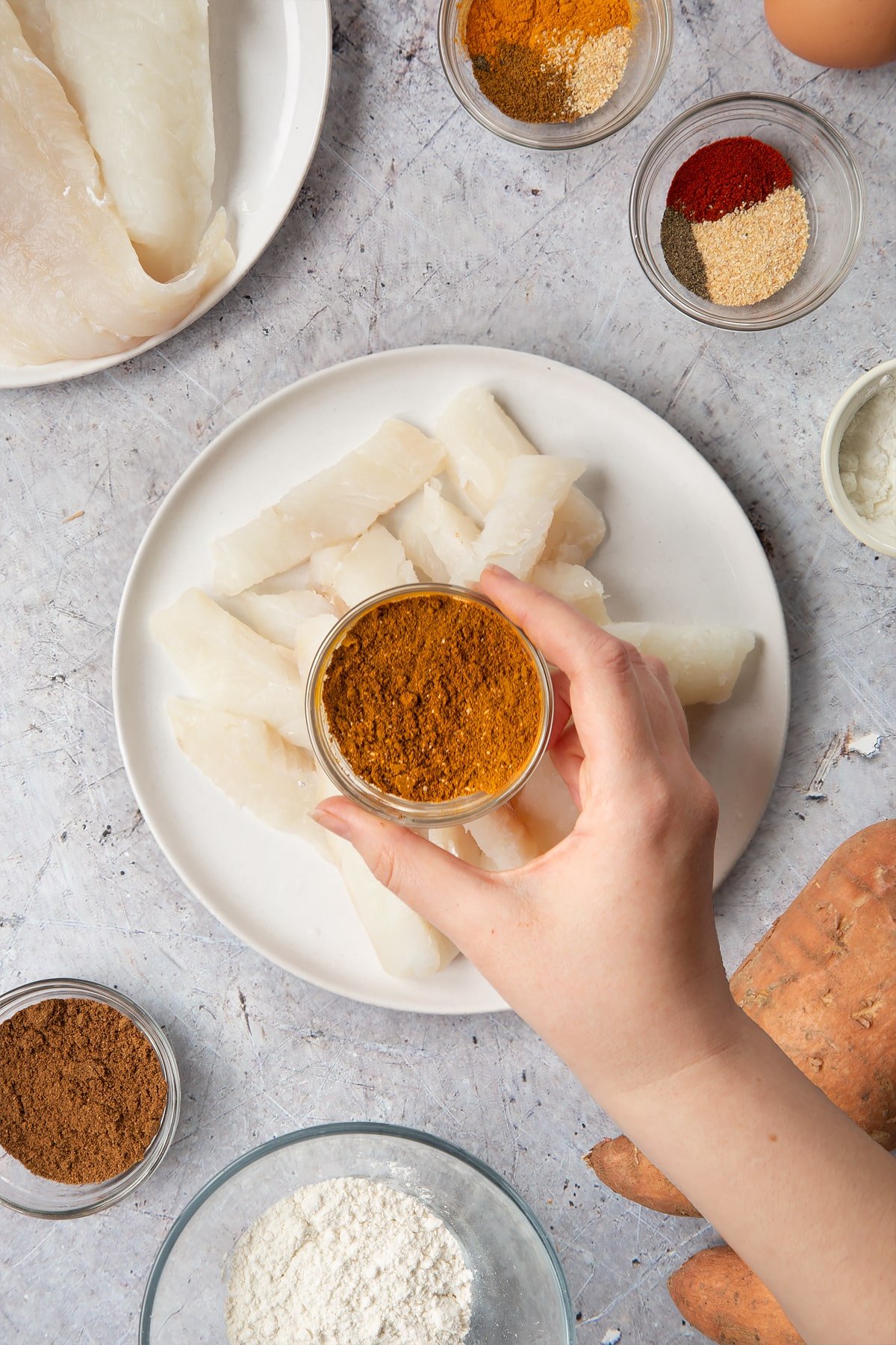 Cod fillets on a white plate, cut into fingers. Above, a hand holds a small bowl containing mixed curry spices. Surrounding the plate is ingredients for spicy fish sticks.