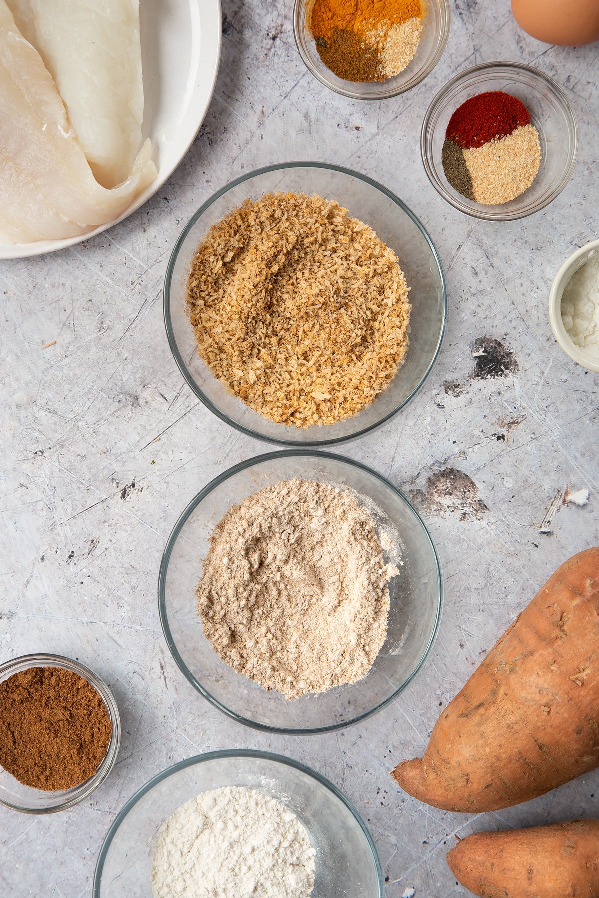 Two bowls, one containing breadcrumbs mixed with spices and another containing flour mixed with spices. Surrounding the bowls is ingredients for spicy fish sticks.