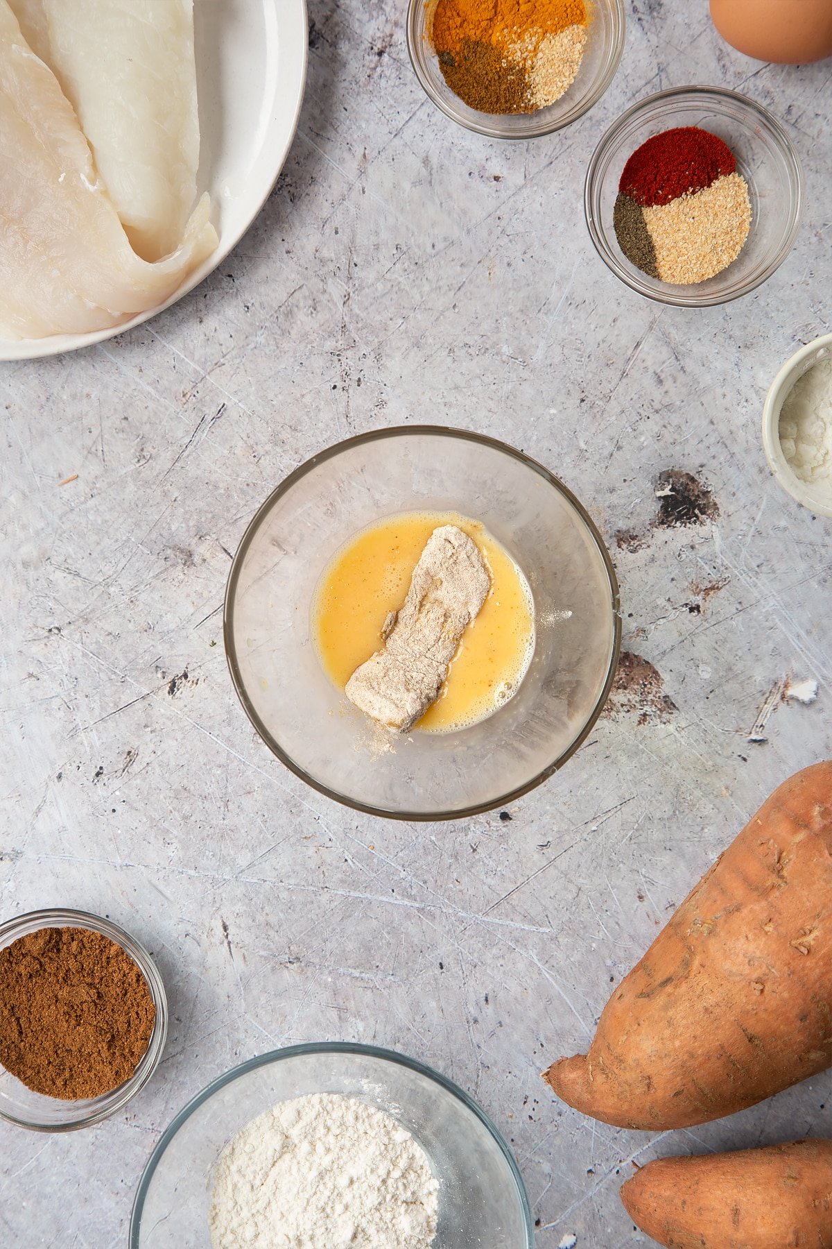 A bowl containing beaten egg, with a piece of flour-coated cod dipped in it. Surrounding the bowl is ingredients for spicy fish sticks.